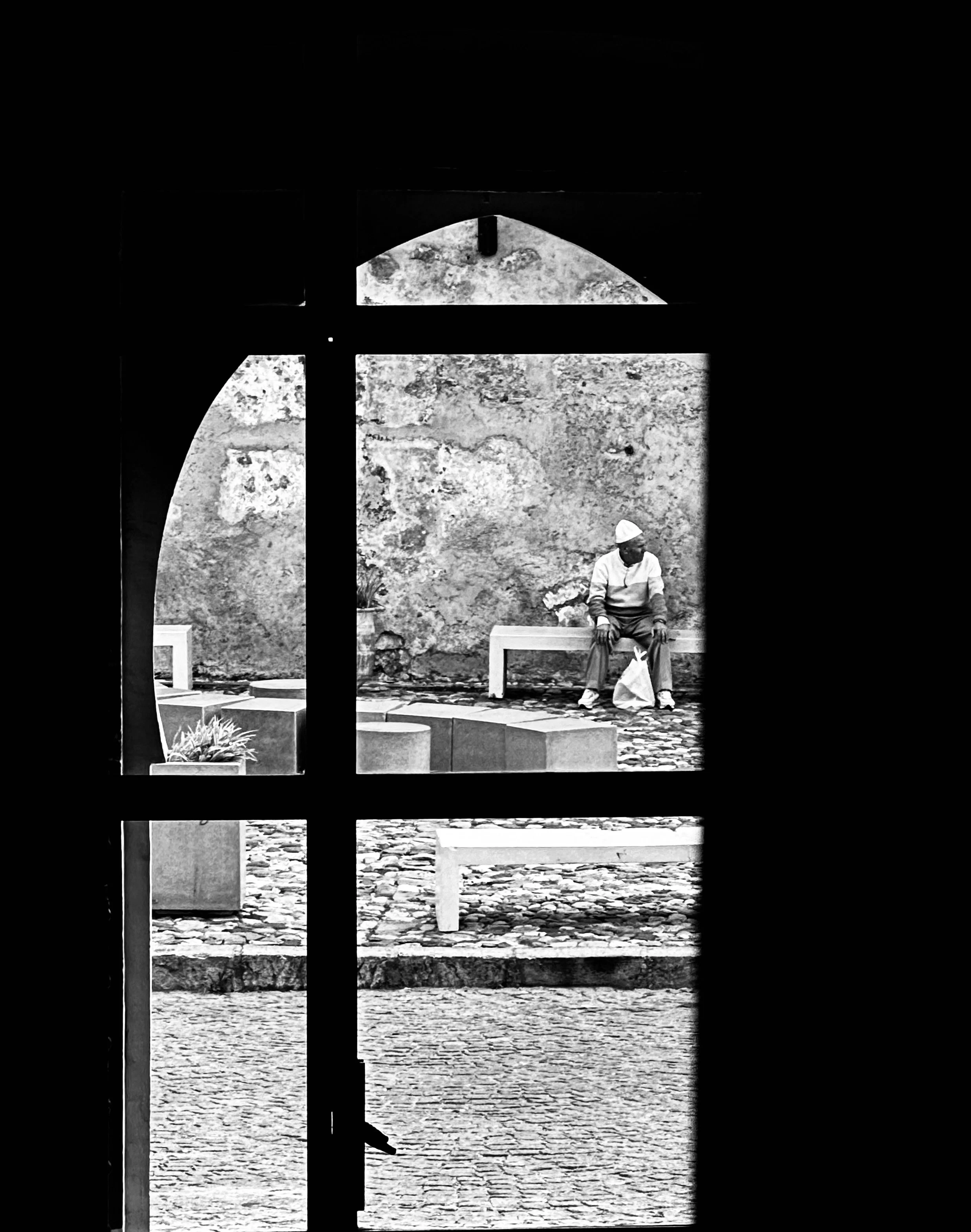 Man sitting on a bench outside, viewed through a window with a cross-shaped frame, with an aged stone wall in the background.