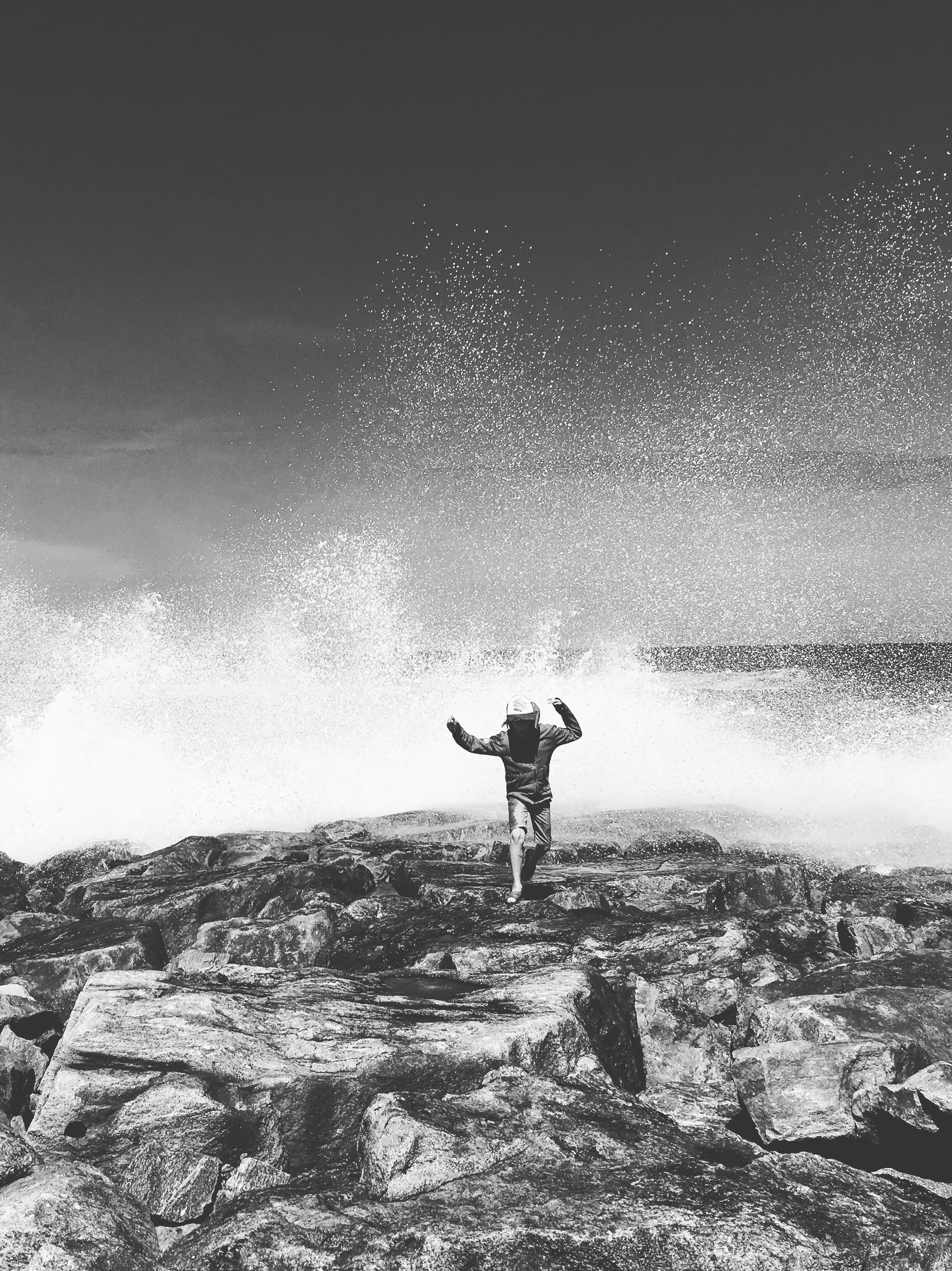 A child running on rocks near the ocean, with waves crashing behind them and spraying water into the air.