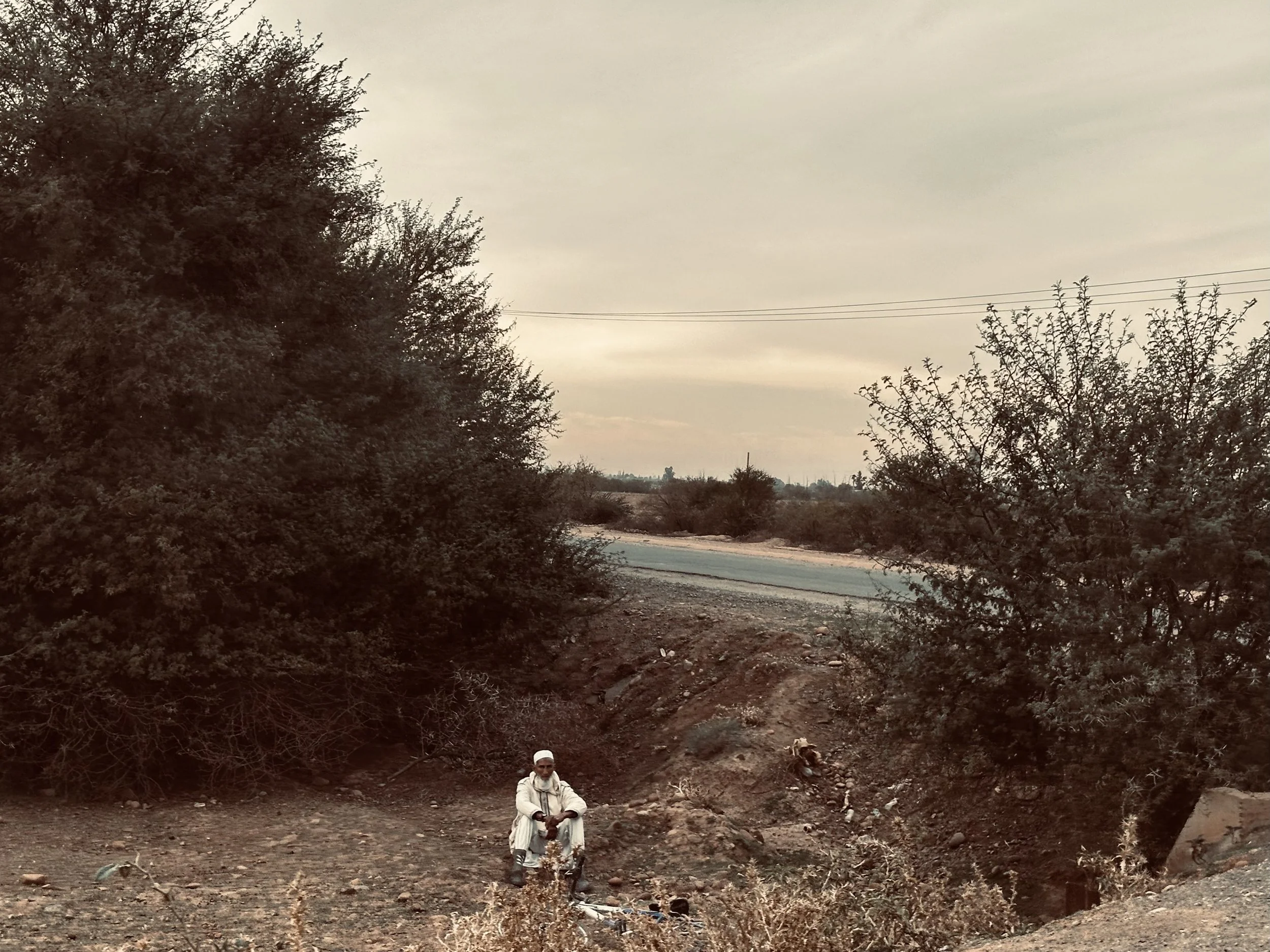 A man wearing traditional white clothing and a white cap sitting on a small bike in a rural area with sparse bushes and trees, dirt ground, a paved road in the background, and an overcast sky.