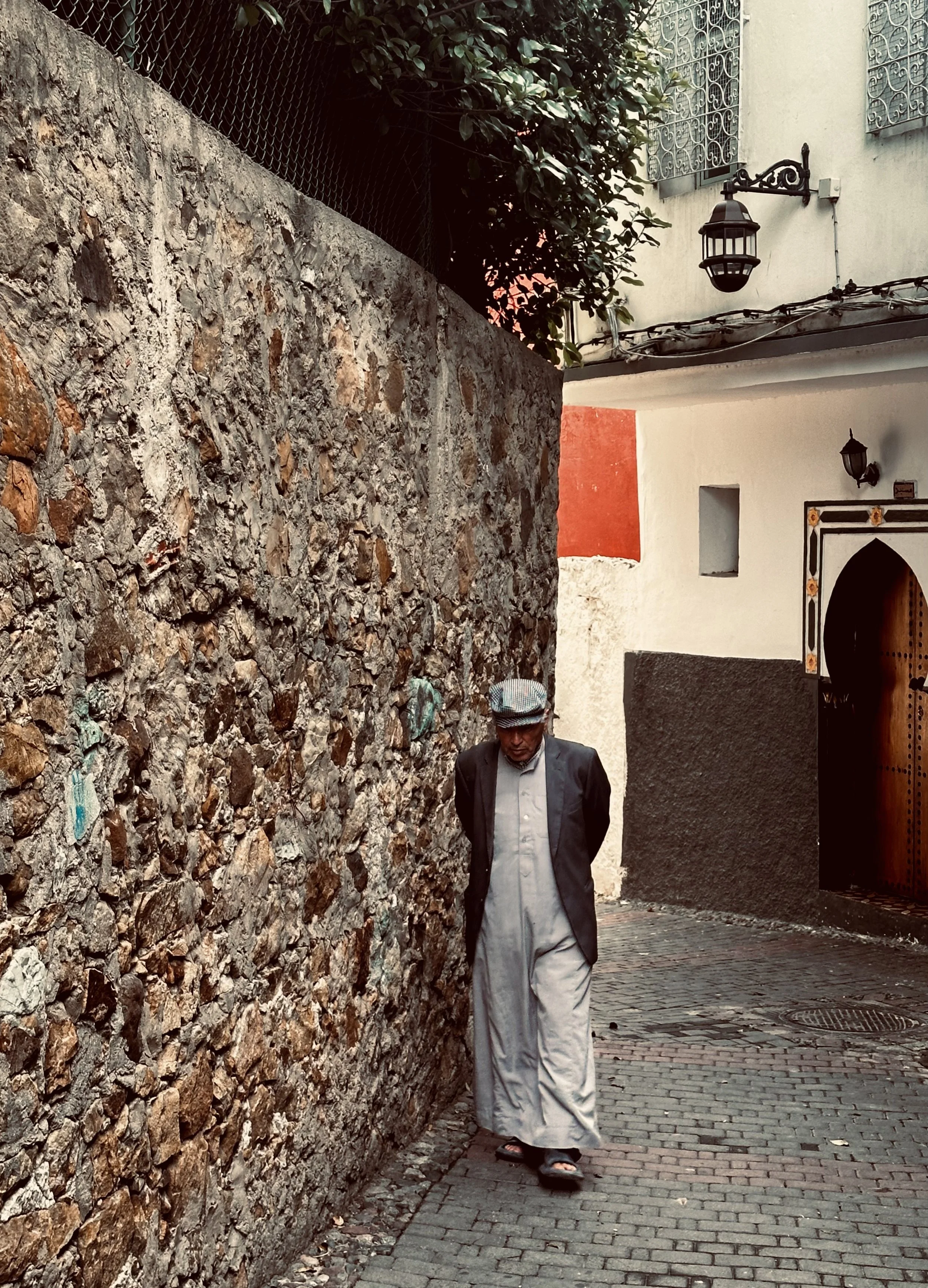 A man wearing traditional clothing, including a cap, walking on a narrow cobblestone street next to a stone wall in an older city area.