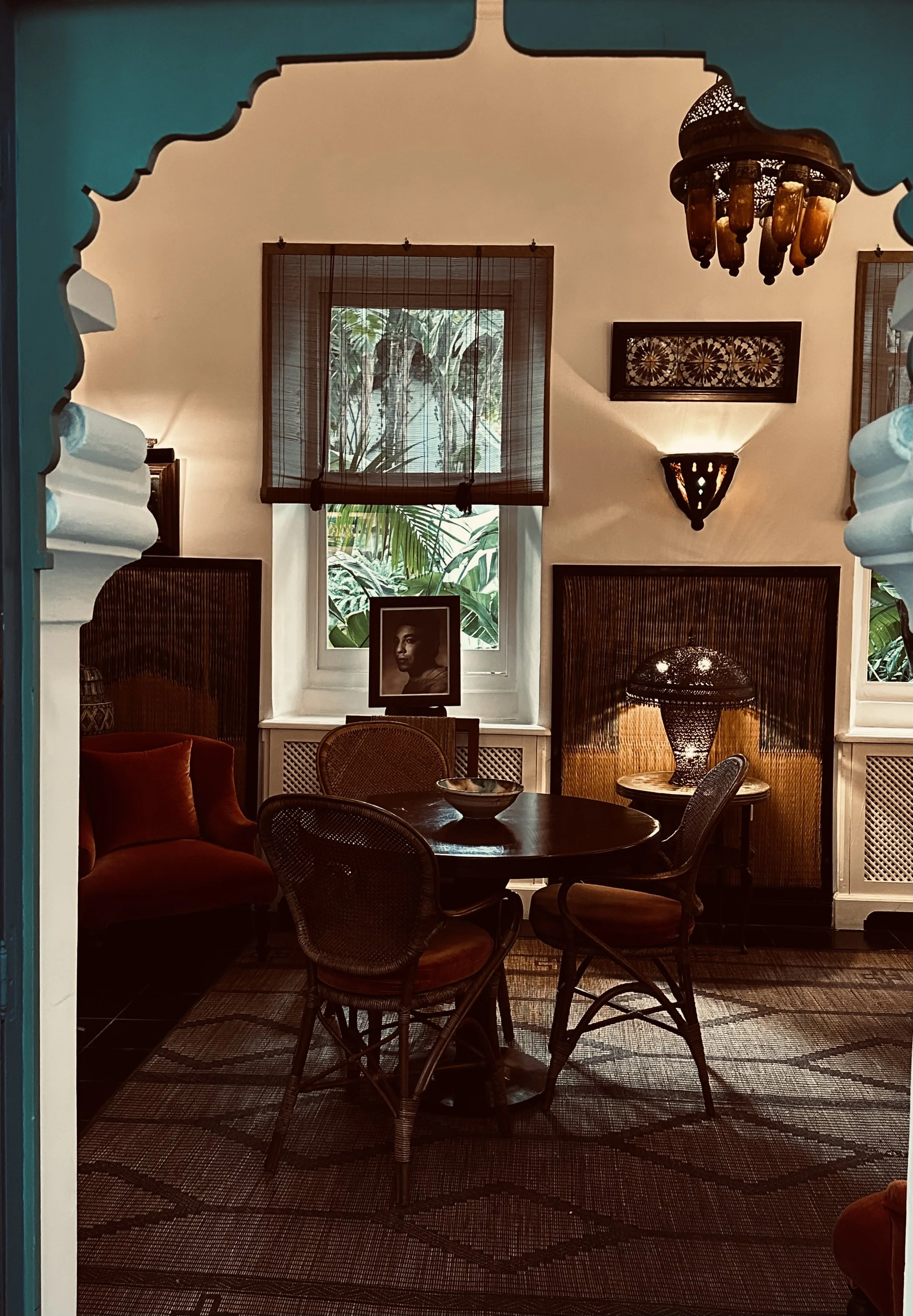 A cozy, vintage-style living room with a round table surrounded by four wicker chairs, a velvet armchair, and eclectic lighting fixtures. The room features two windows with wooden blinds, a framed black and white portrait on the window ledge, and tro