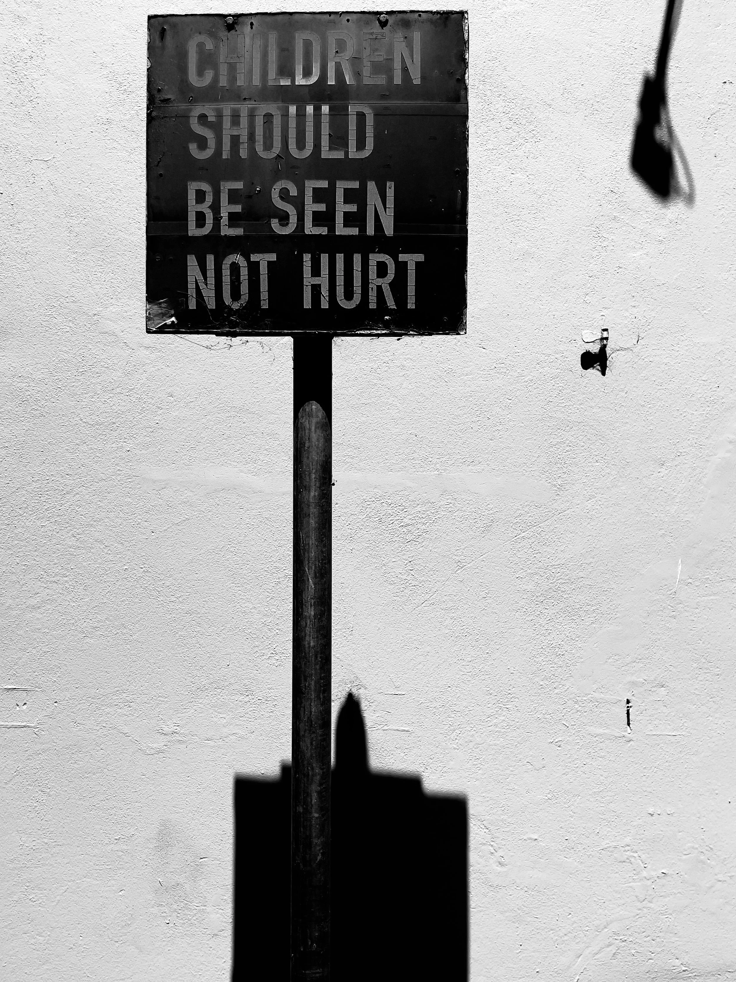 A black sign with the words "Children should be seen not hurt" in faded capital letters, mounted on a pole against a plain white wall.