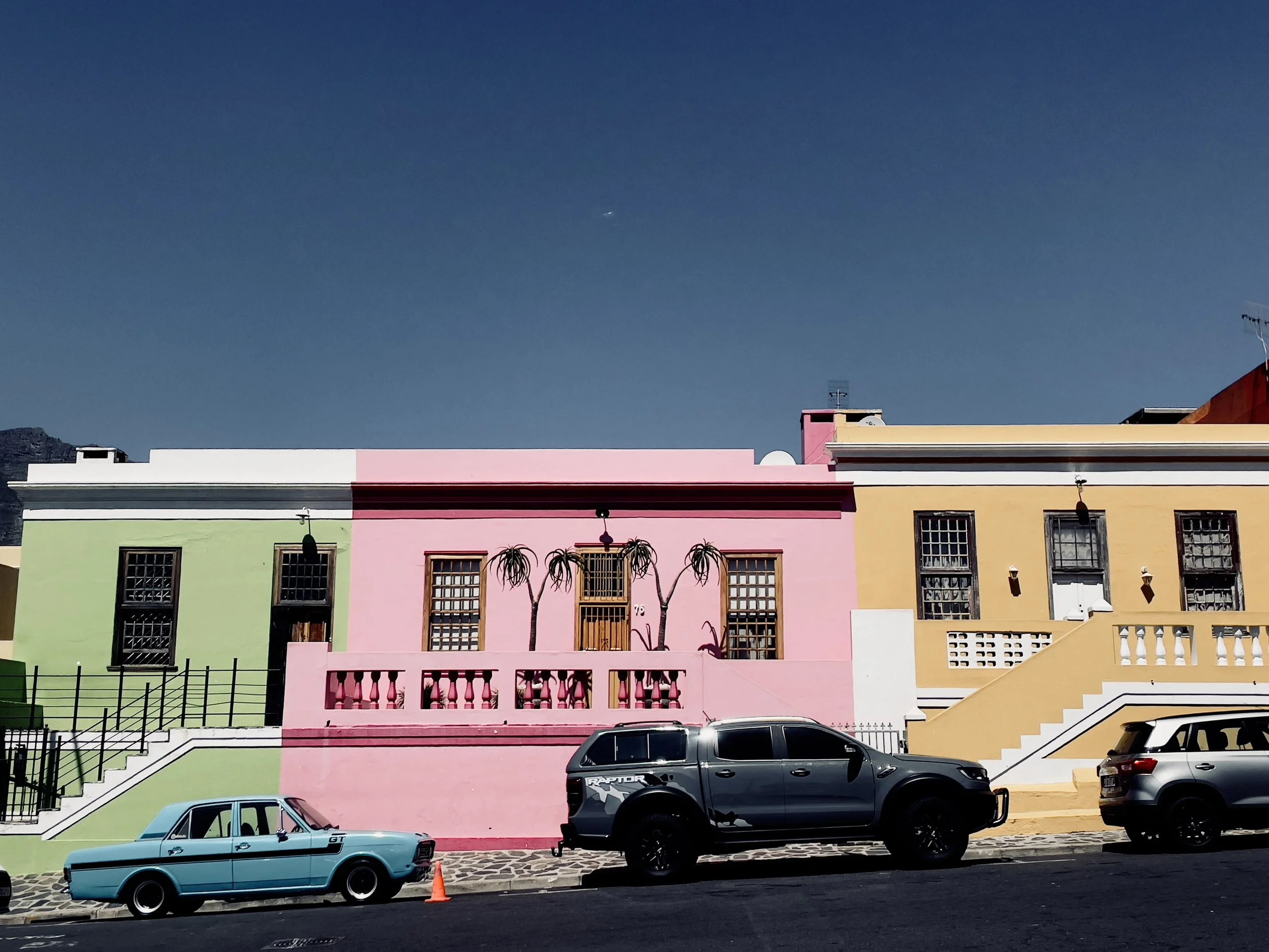 Colorful row of houses painted in green, pink, and yellow, with cars parked in front and a clear blue sky above.
