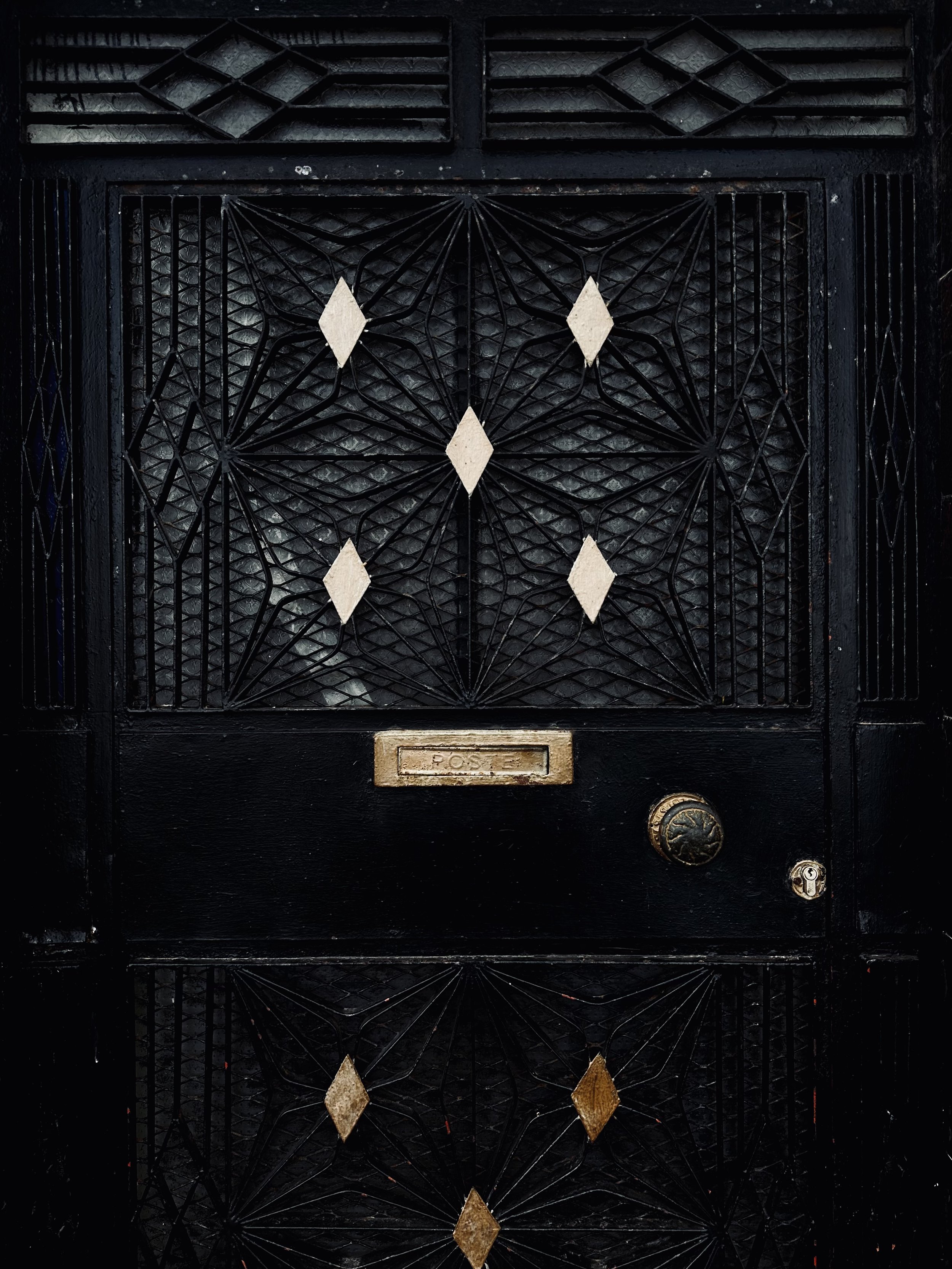 Old black mailbox with decorative metal grille, diamond-shaped embellishments, and a brass lock near the bottom.