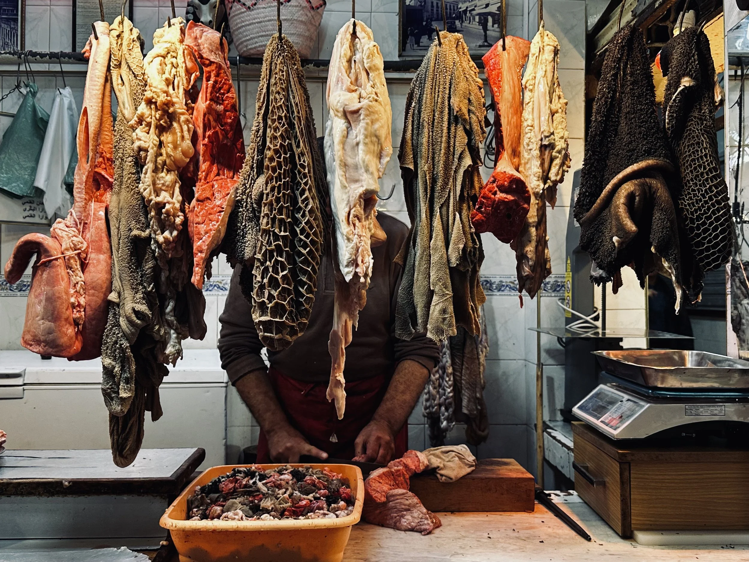A butcher's stall with hanging animal hides and cuts of meat. The person is partially obscured behind the hanging carcasses in a market setting.