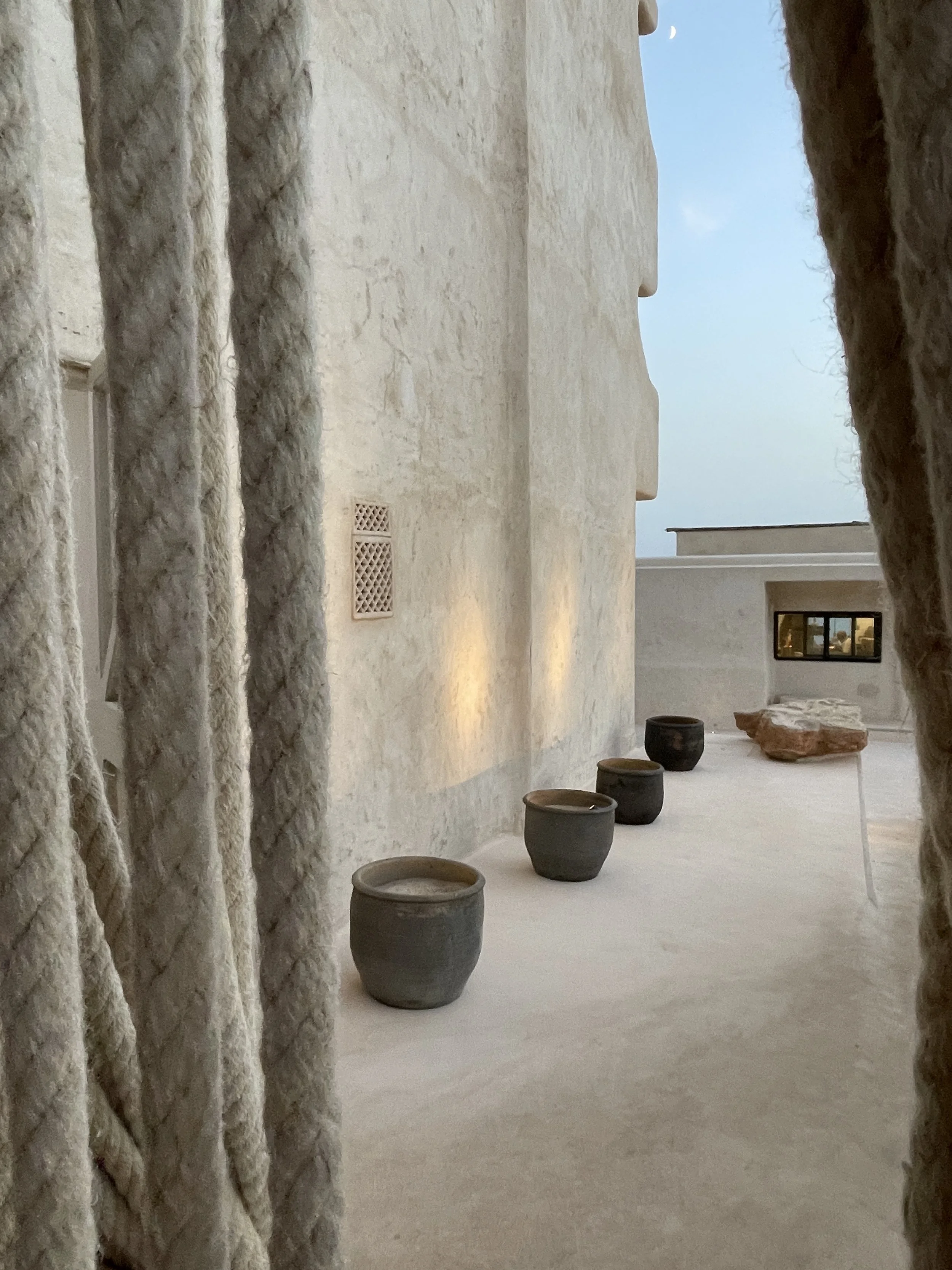 A view of a minimalist outdoor terrace with a textured white wall, pottery planters, and rocks, seen through thick, twisted rope curtains, with a soft evening light.