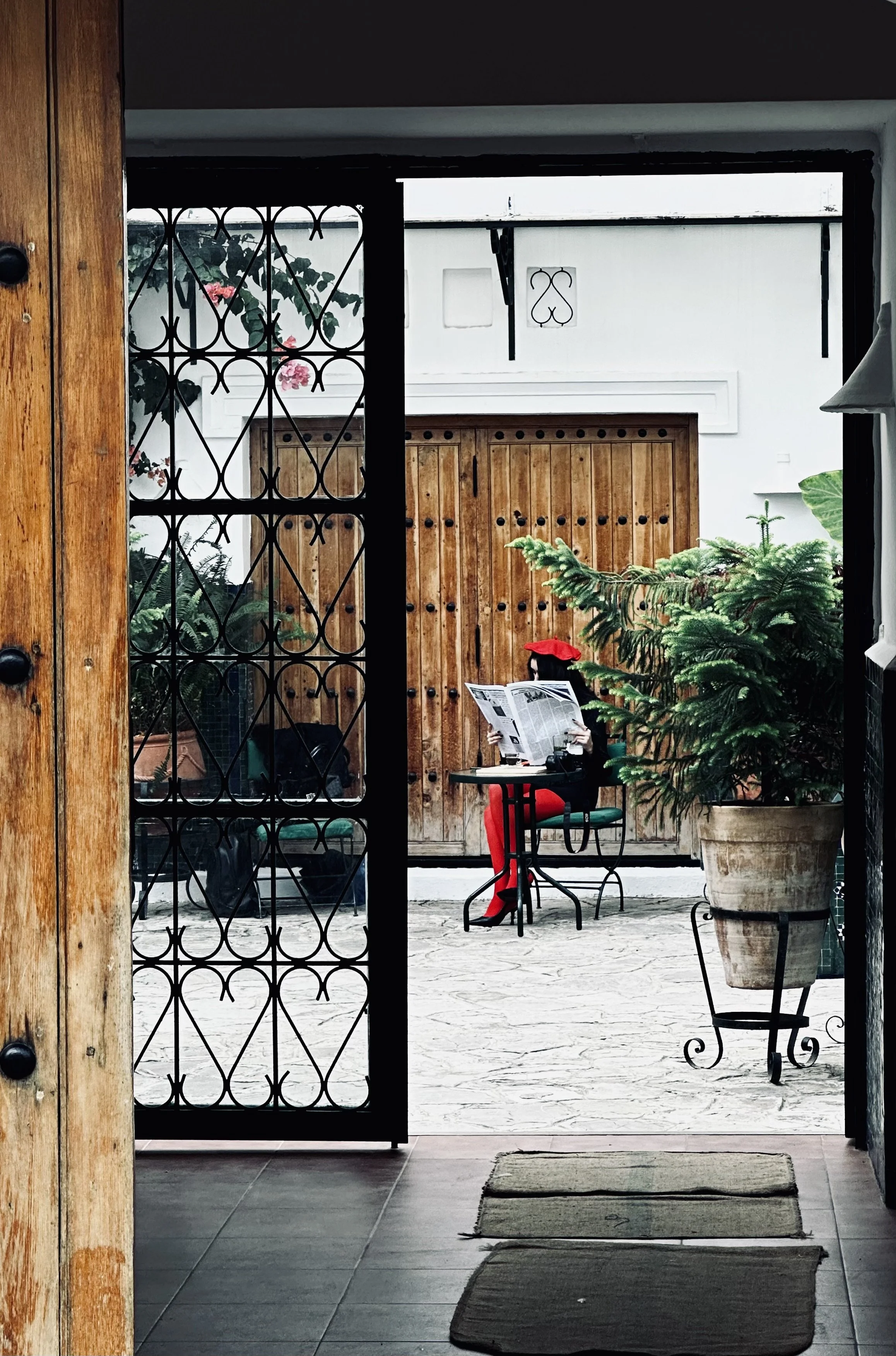 A person wearing red clothing and a red hat is sitting at an outdoor table reading a newspaper, with large potted plants and a wooden door in the background.