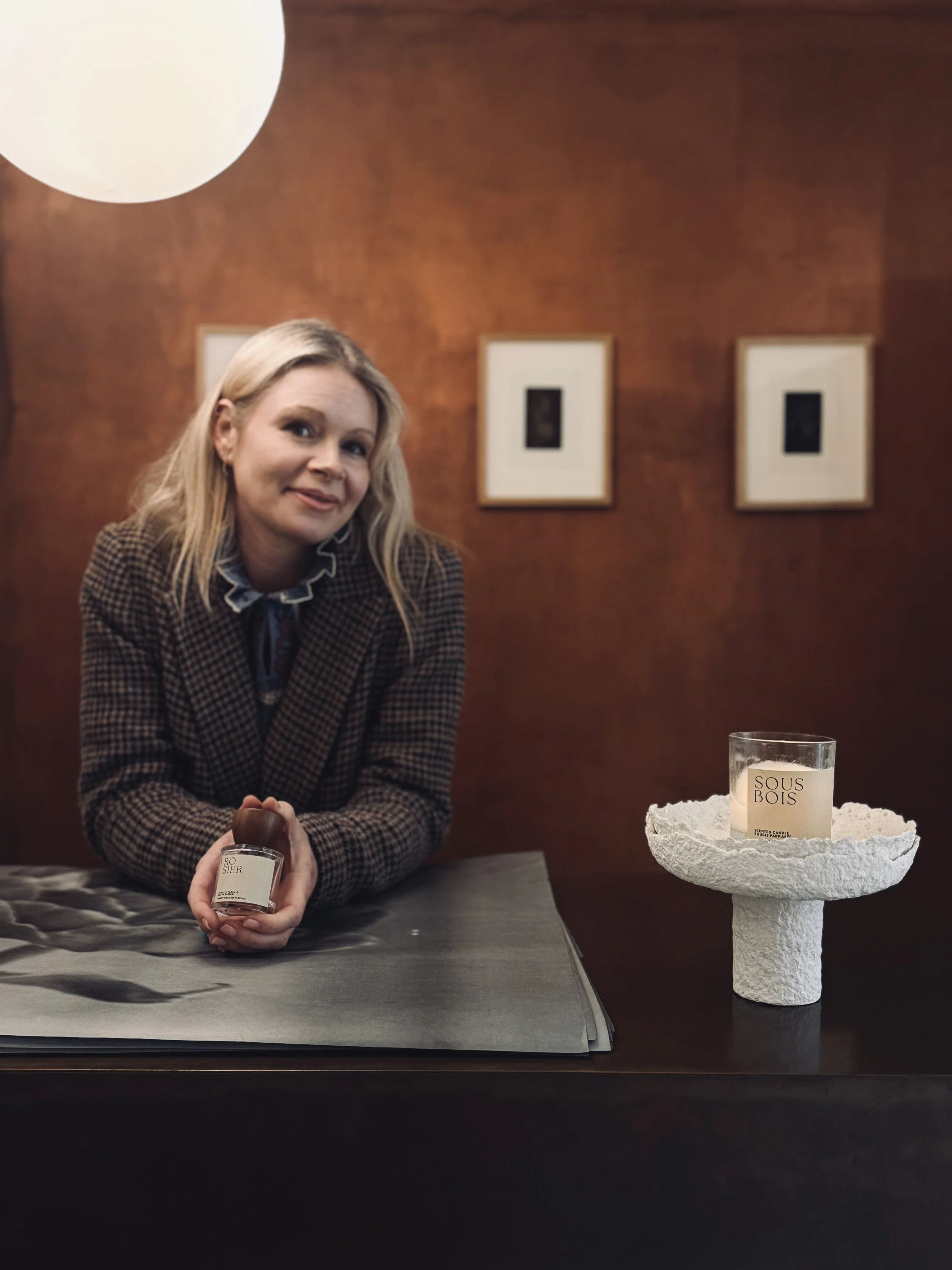 Woman with blonde hair in checkered blazer holding a candle on a table with dark woods background and framed pictures on the wall.