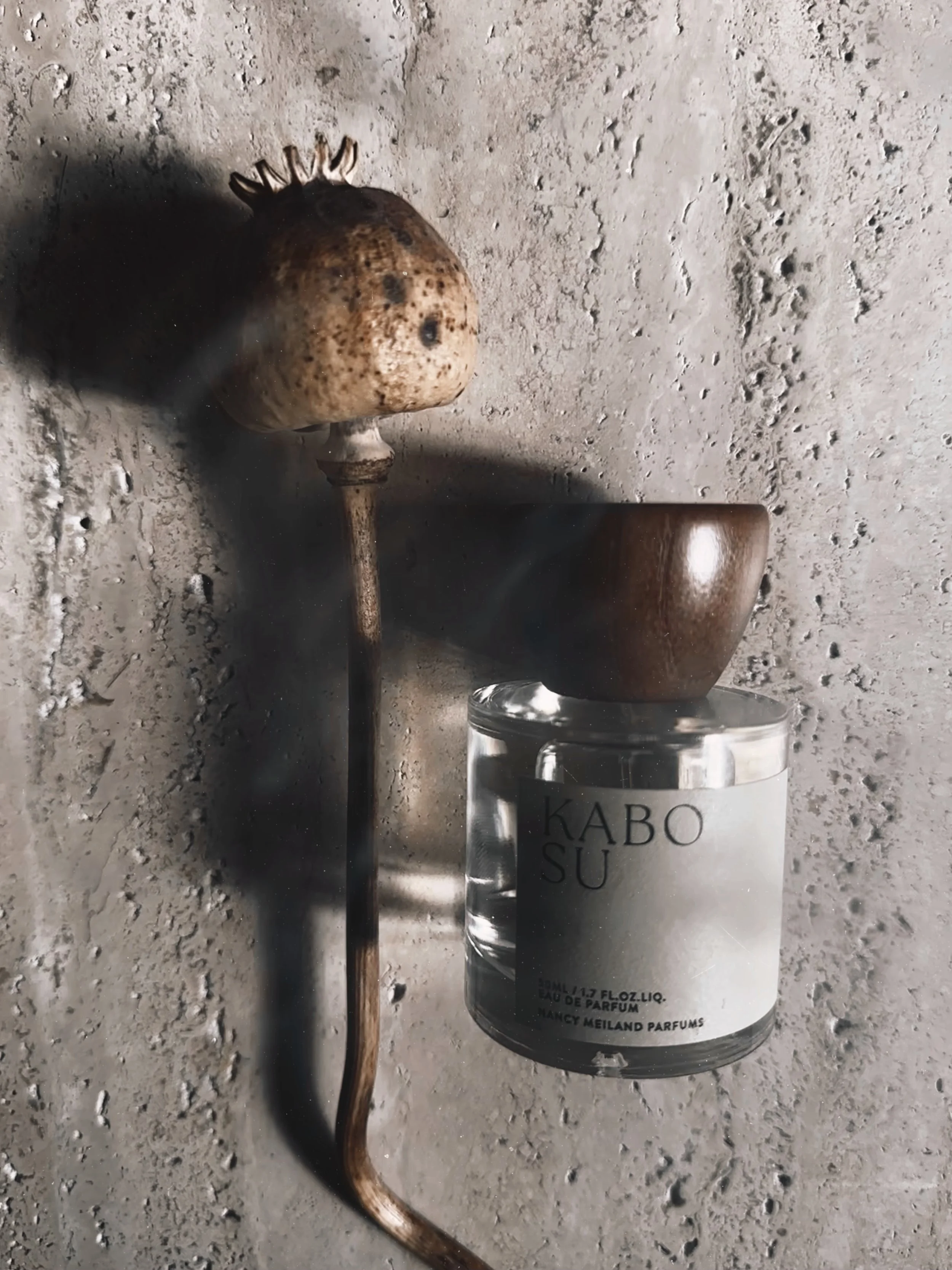 A vintage object resembling an old-fashioned lamp with a bulbous top and long, curved stem, placed on a textured concrete surface next to a patterned bowl and a glass jar labeled 'KABOSU' containing perfume.