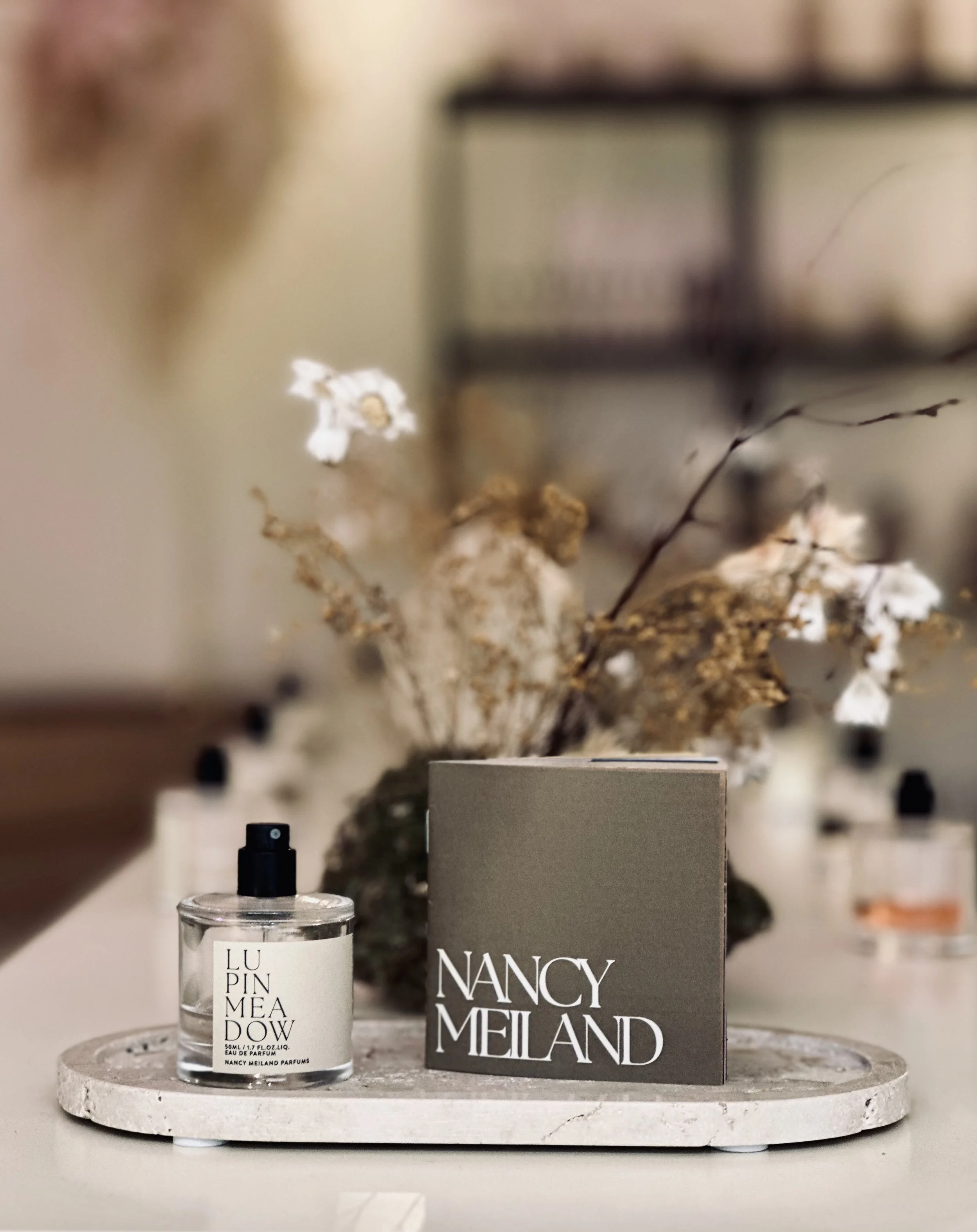 A perfume bottle labeled Nancy Meland with a spray nozzle, a gray box with Nancy Meland written in white, and a decorative arrangement of dried flowers in the background on a white marble tray.