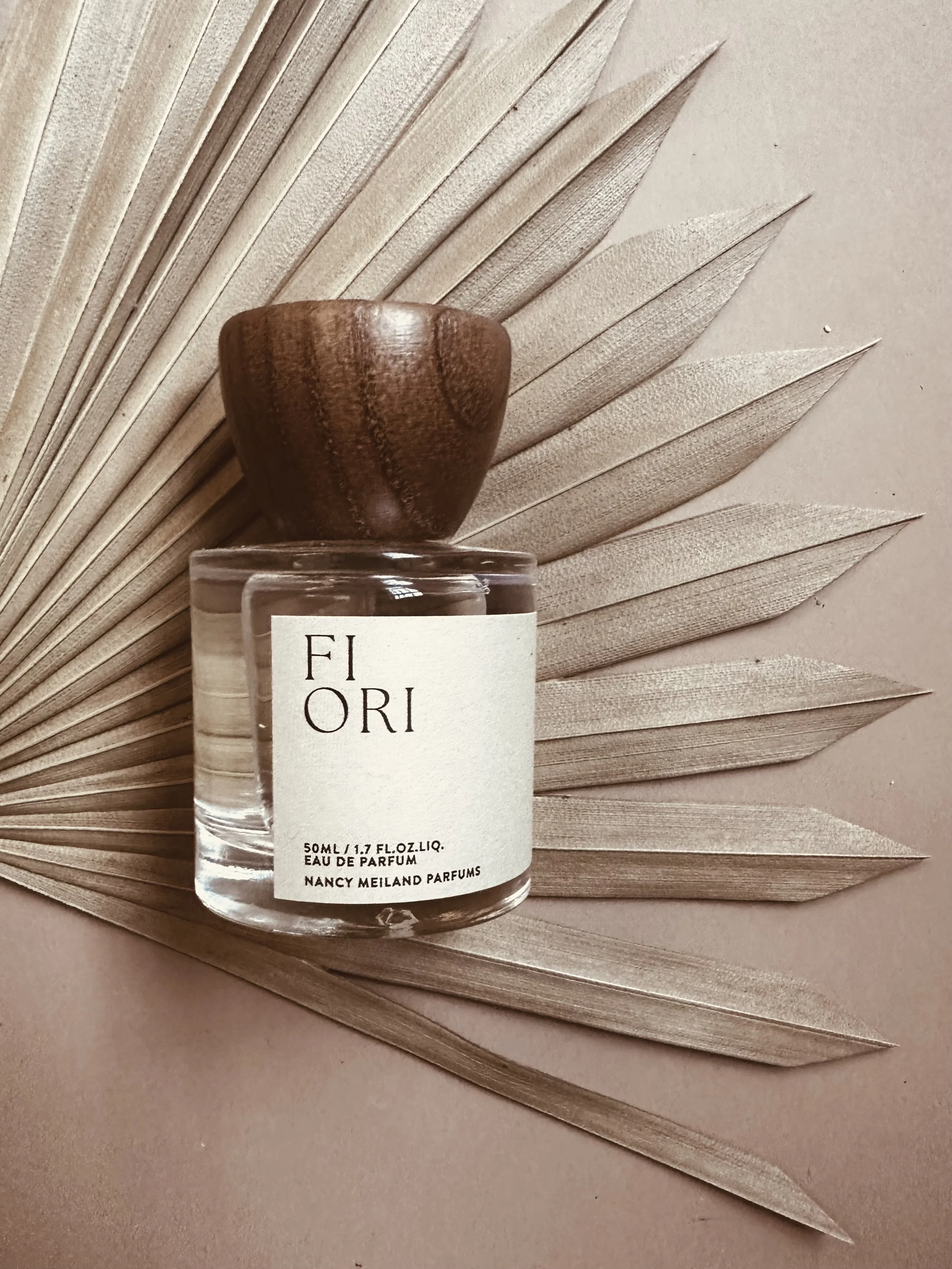 A glass perfume bottle with a white label and a wooden cap, placed on a neutral-colored surface with a dried palm leaf in the background.