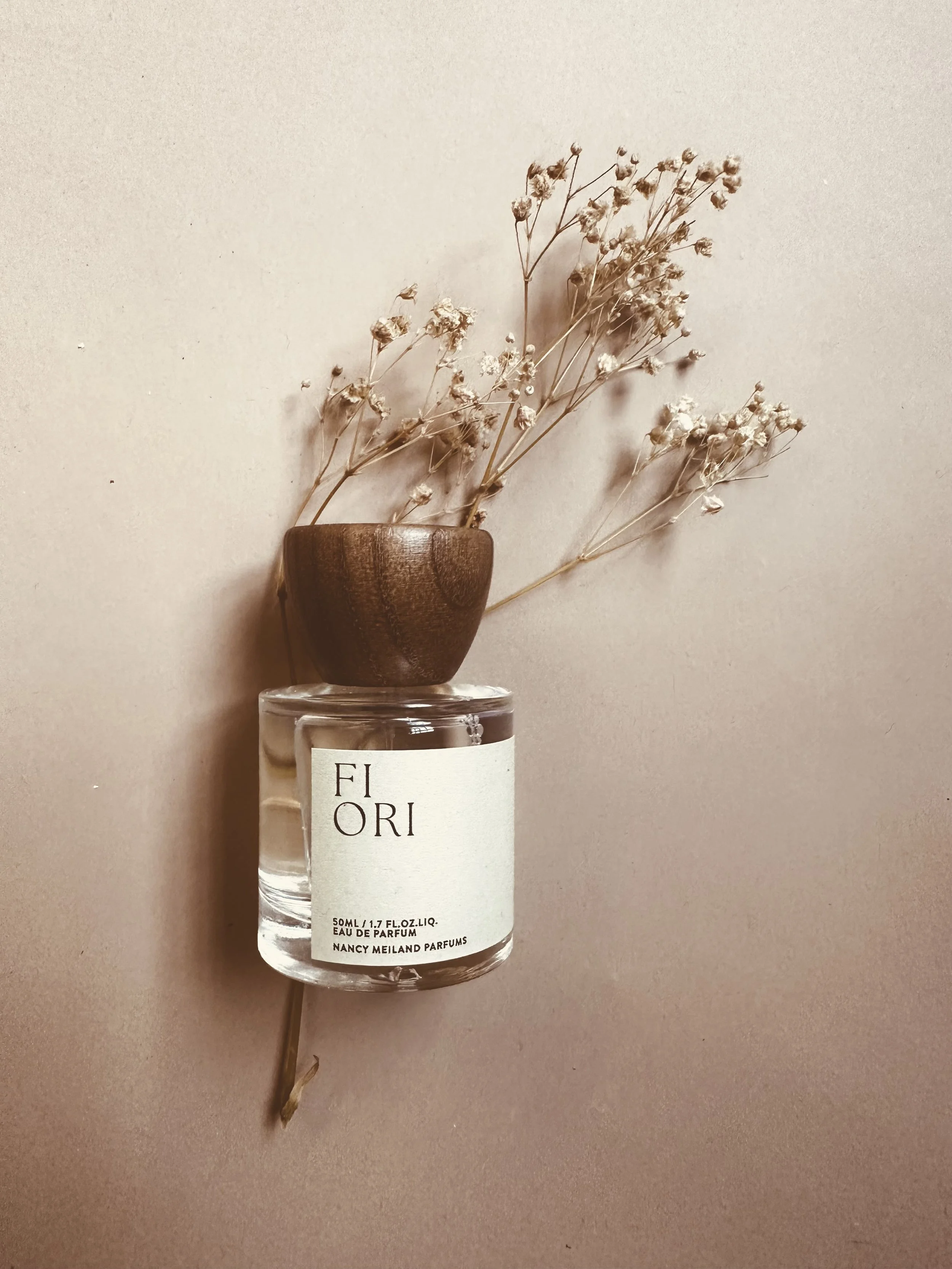 A perfume bottle with a wooden cap, with dried flowers placed in it, on top of another perfume bottle, against a plain background.