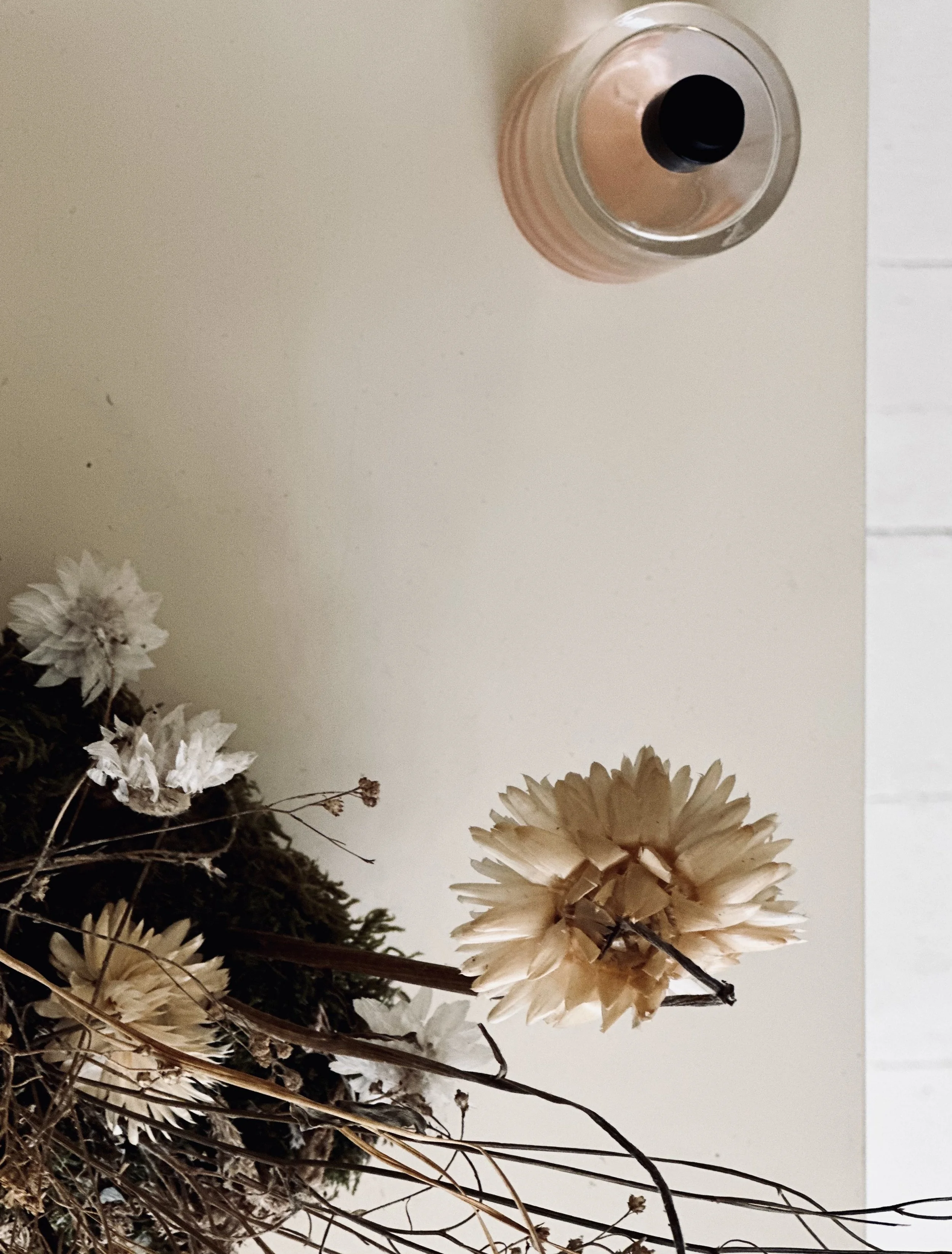 A top view of a beige table with a beige candle with a black wick, dried flowers, and foam flower arrangement.