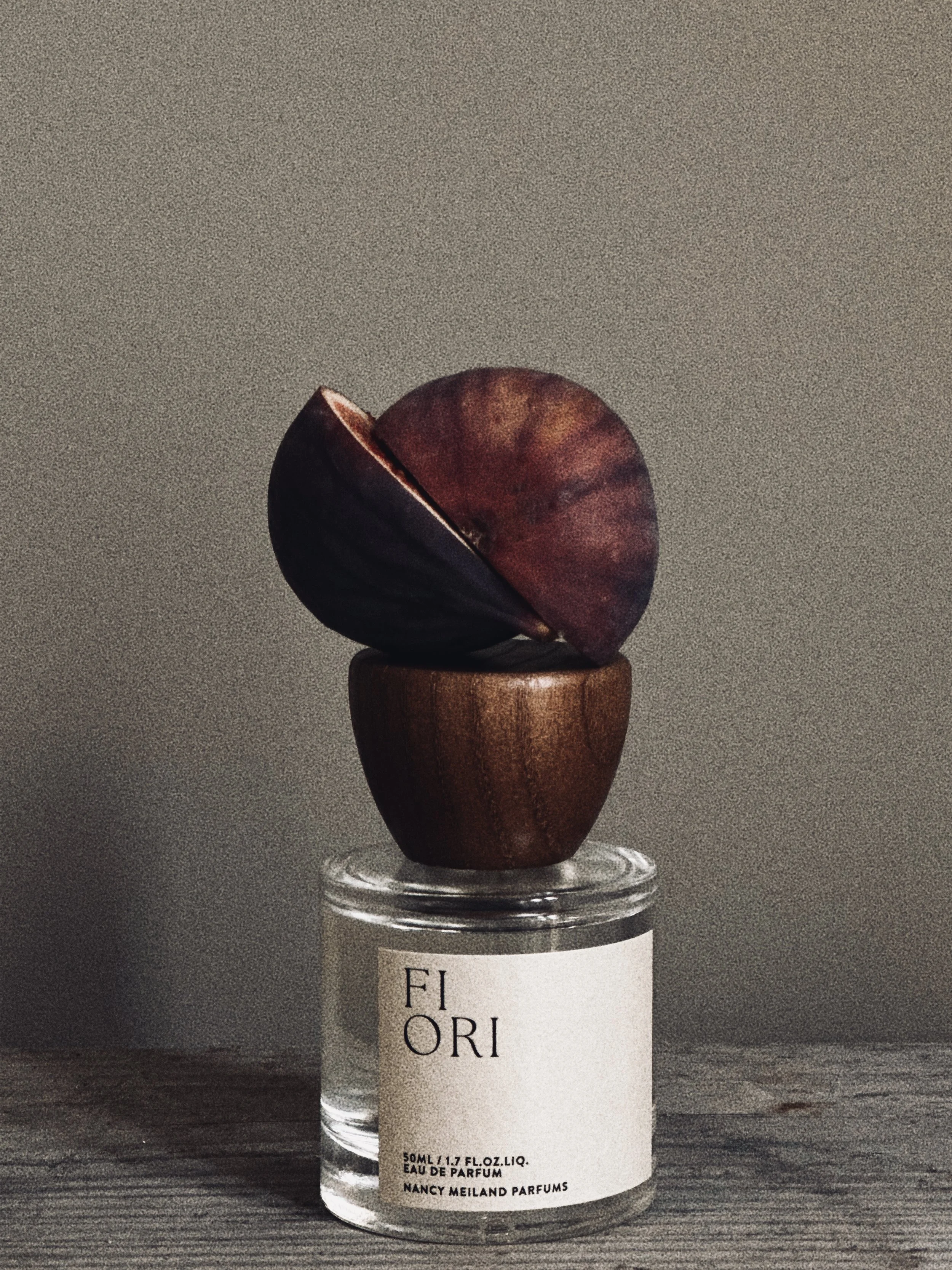 A perfume bottle labeled 'FIORI' with a wooden cap, topped with a hollowed-out purple and reddish fruit or seed, placed on a textured surface against a neutral background.
