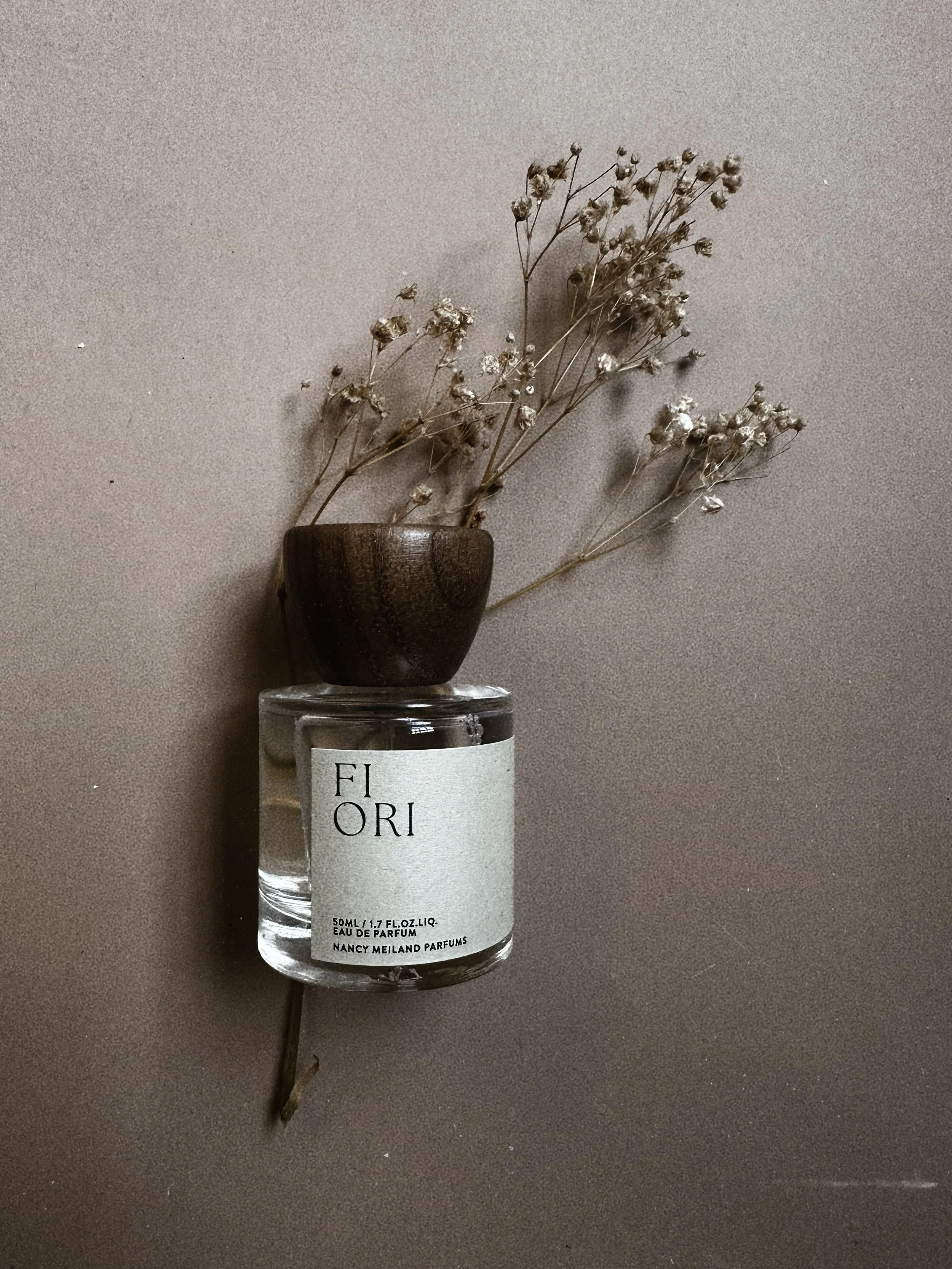 Dried flowers in a small wooden cup placed on top of a perfume bottle labeled 'Fiori' with a plain background.