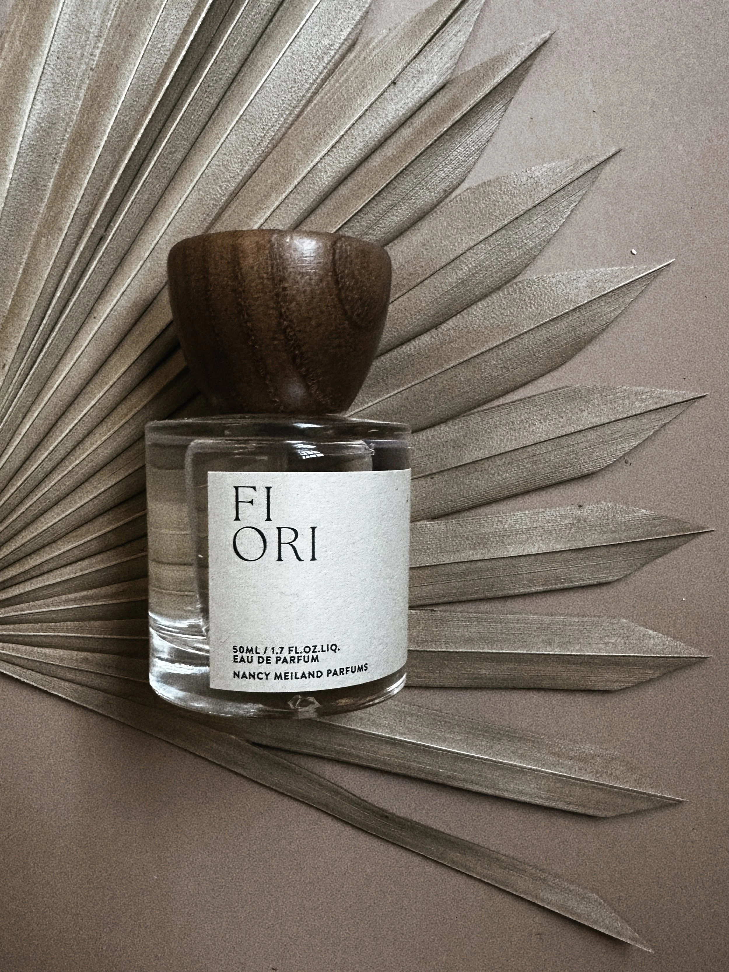 A perfume bottle labeled 'Fior' with a wooden cap, placed on a metallic palm leaf-shaped decorative piece with a soft pink background.