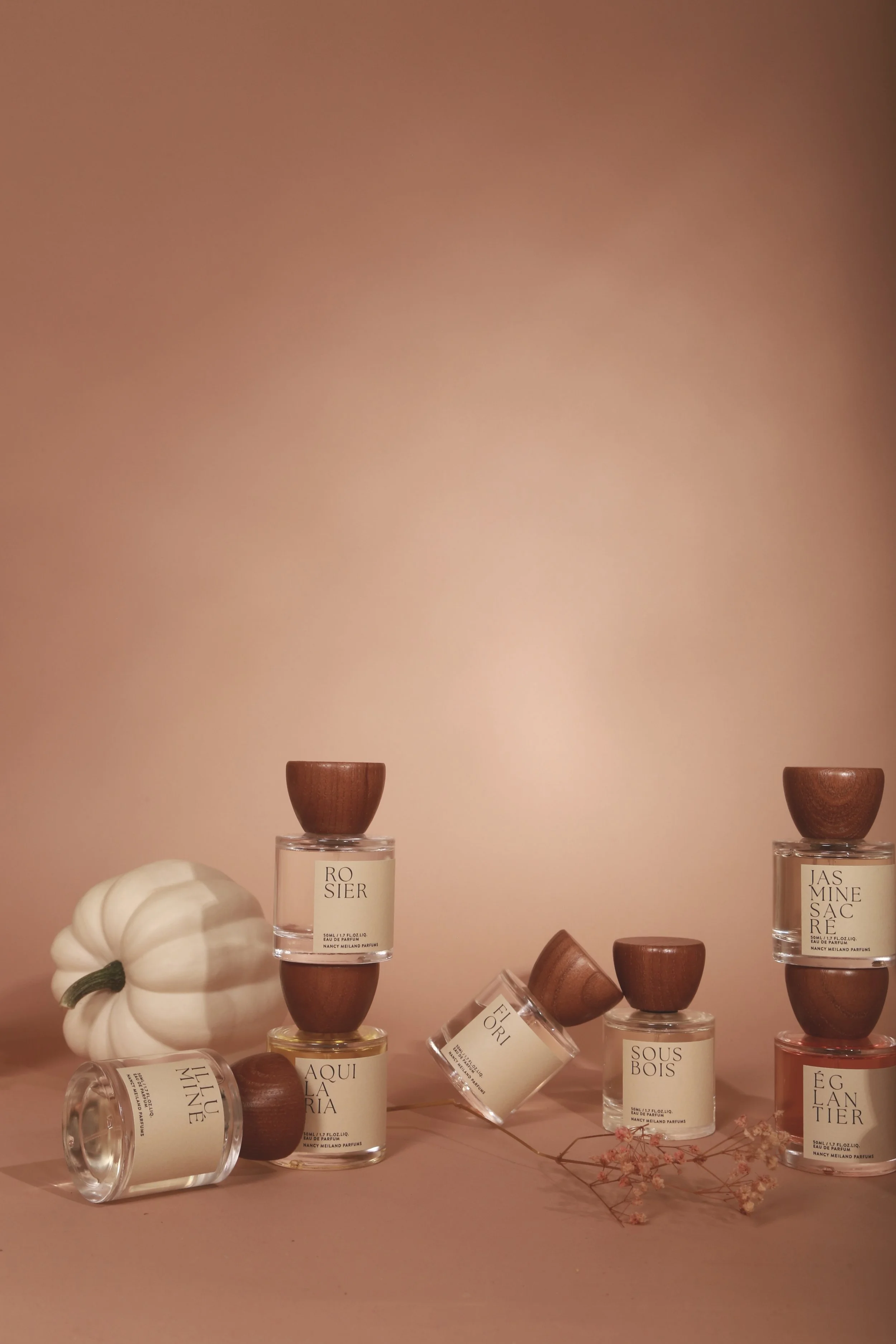 Display of several scented candles and a white pumpkin against a pink background, with some small flowers.