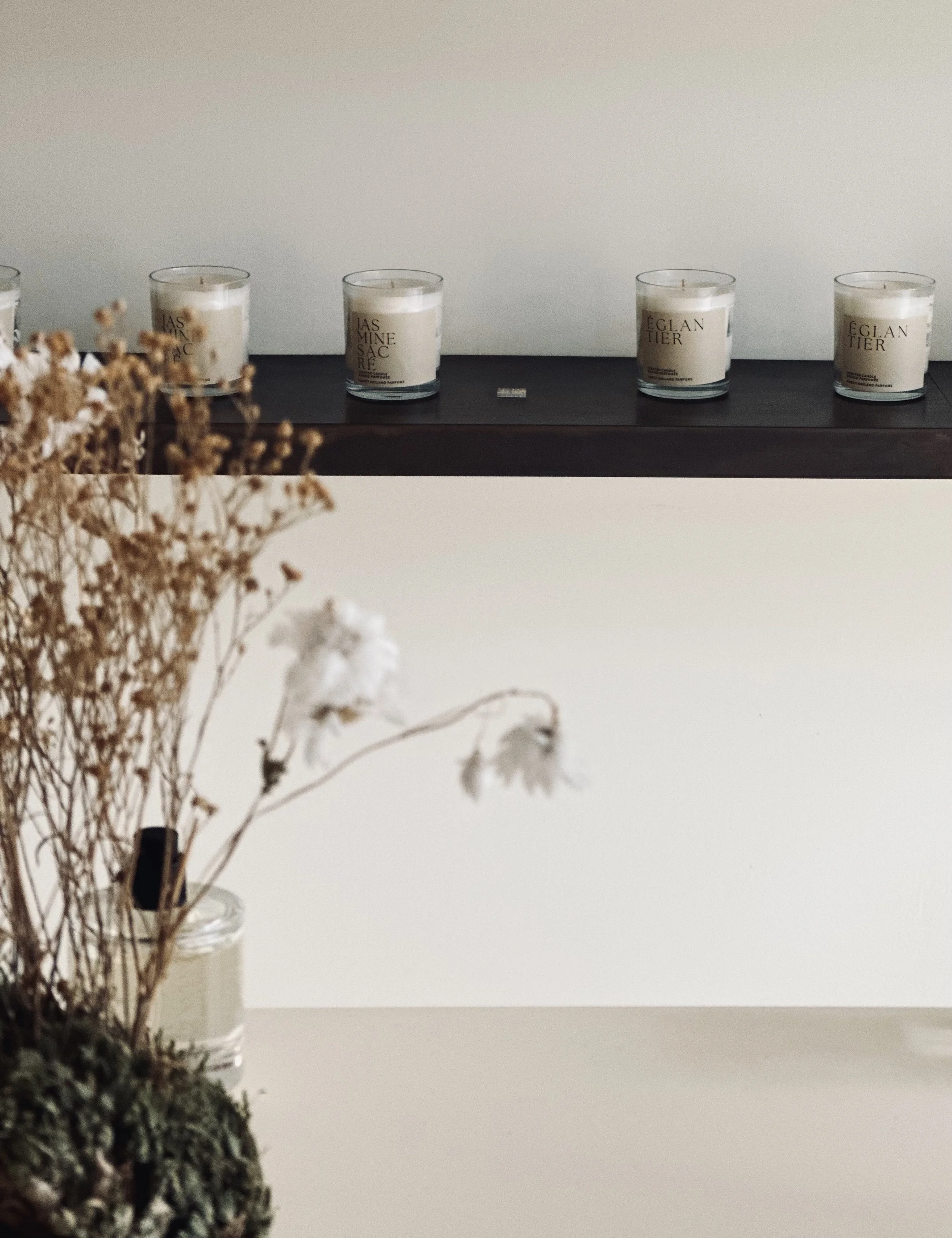 A white wall with a dark wooden shelf holding four white candles in glass containers, with dried flowers in a glass jar in the foreground.