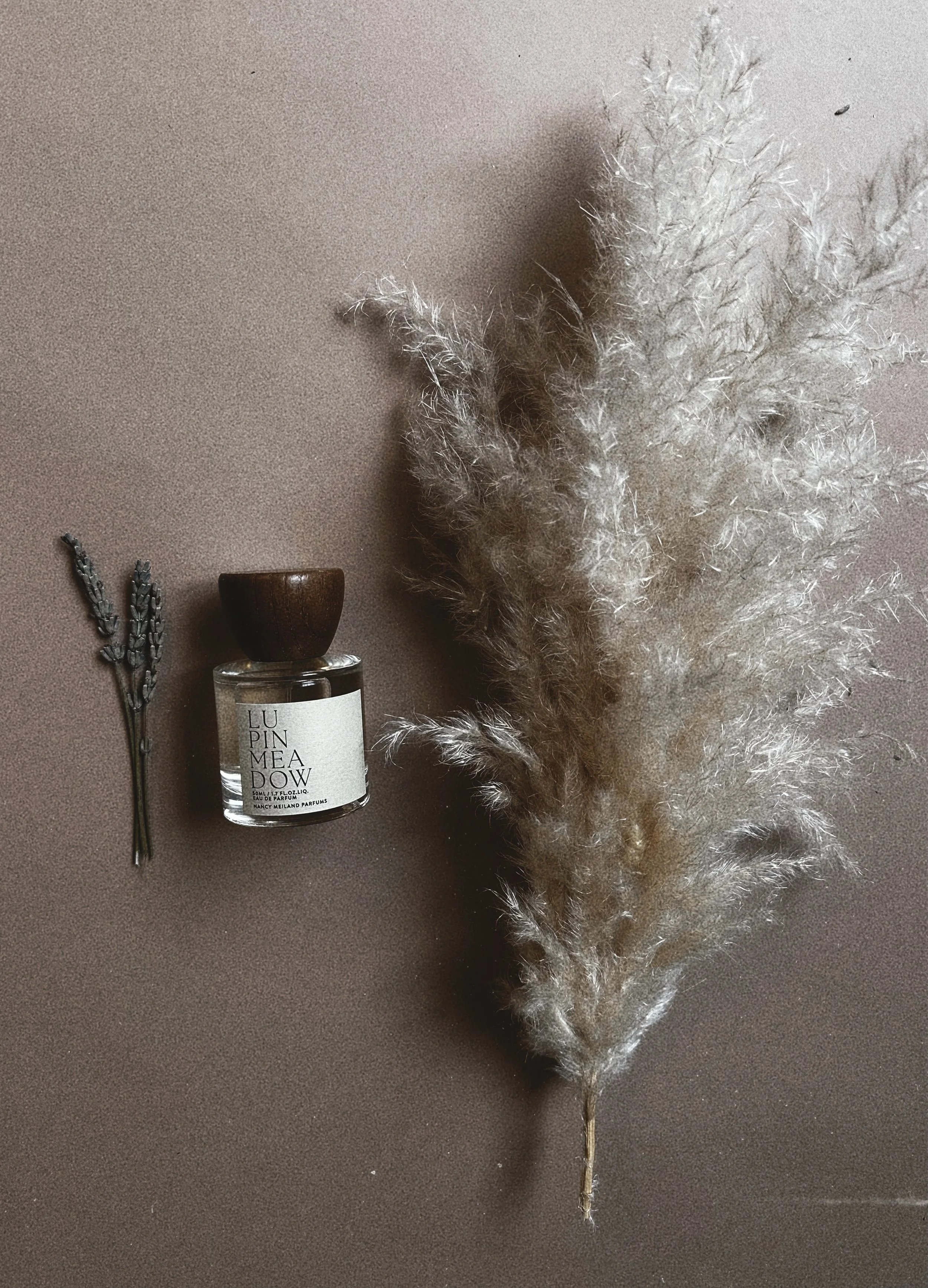 A small bunch of lavender, a bottle of perfume labeled 'LUMINEMADEW,' and a large fluffy dried pampas grass stem are arranged on a neutral-colored surface.