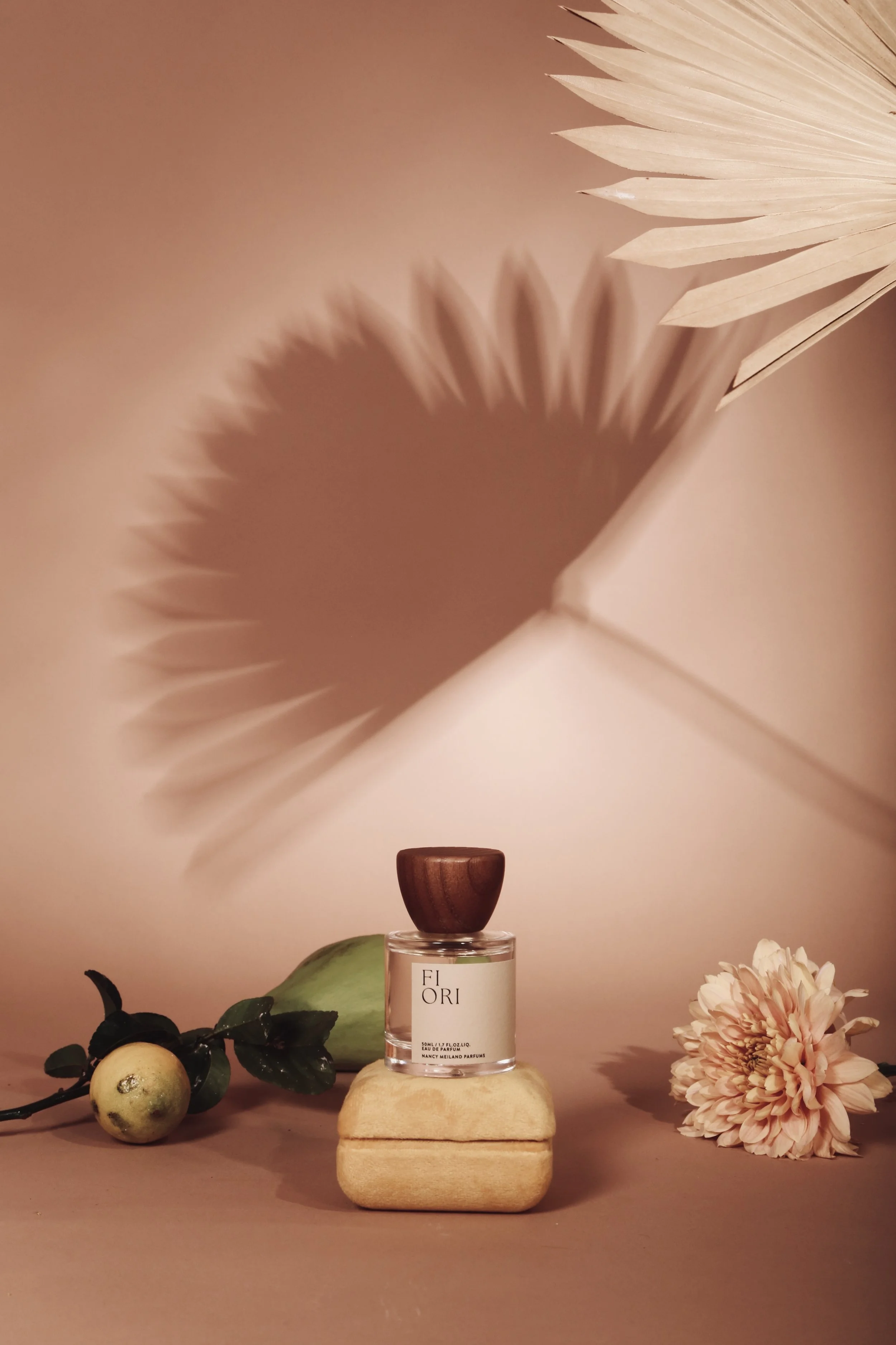 A perfume bottle labeled 'FIORI' on a beige surface, surrounded by a flower, a fruit, and shadow of a leaf on the wall.