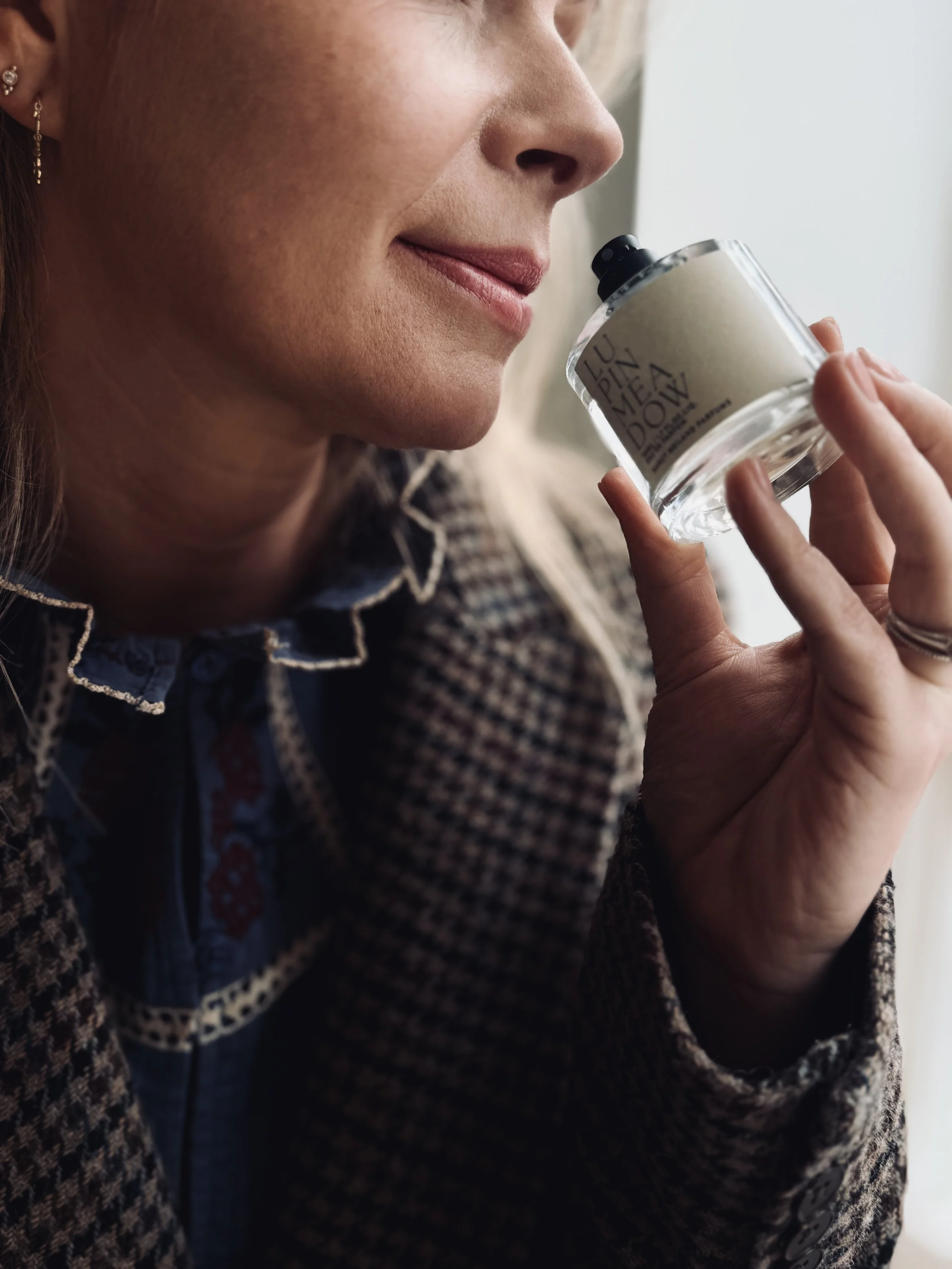 Close-up of a woman holding a small glass container of cream or balm with a black screw cap, near her nose, with a window in the background.