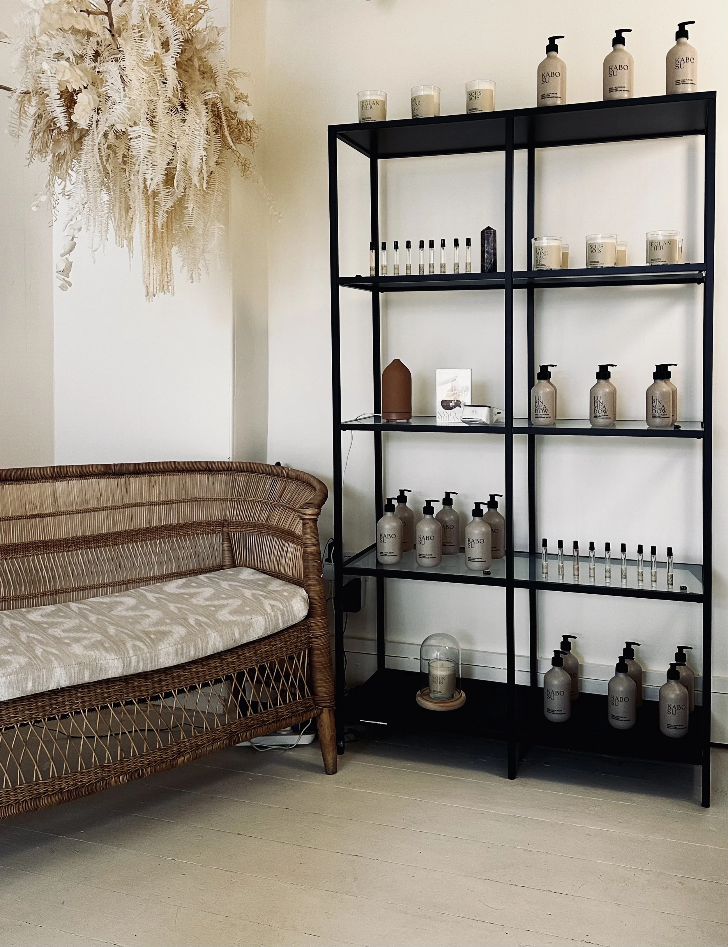 Interior of a room with a rattan sofa, a black metal shelving unit holding bottles and candles, a large dried floral arrangement hanging on the wall, and light-colored wooden flooring.