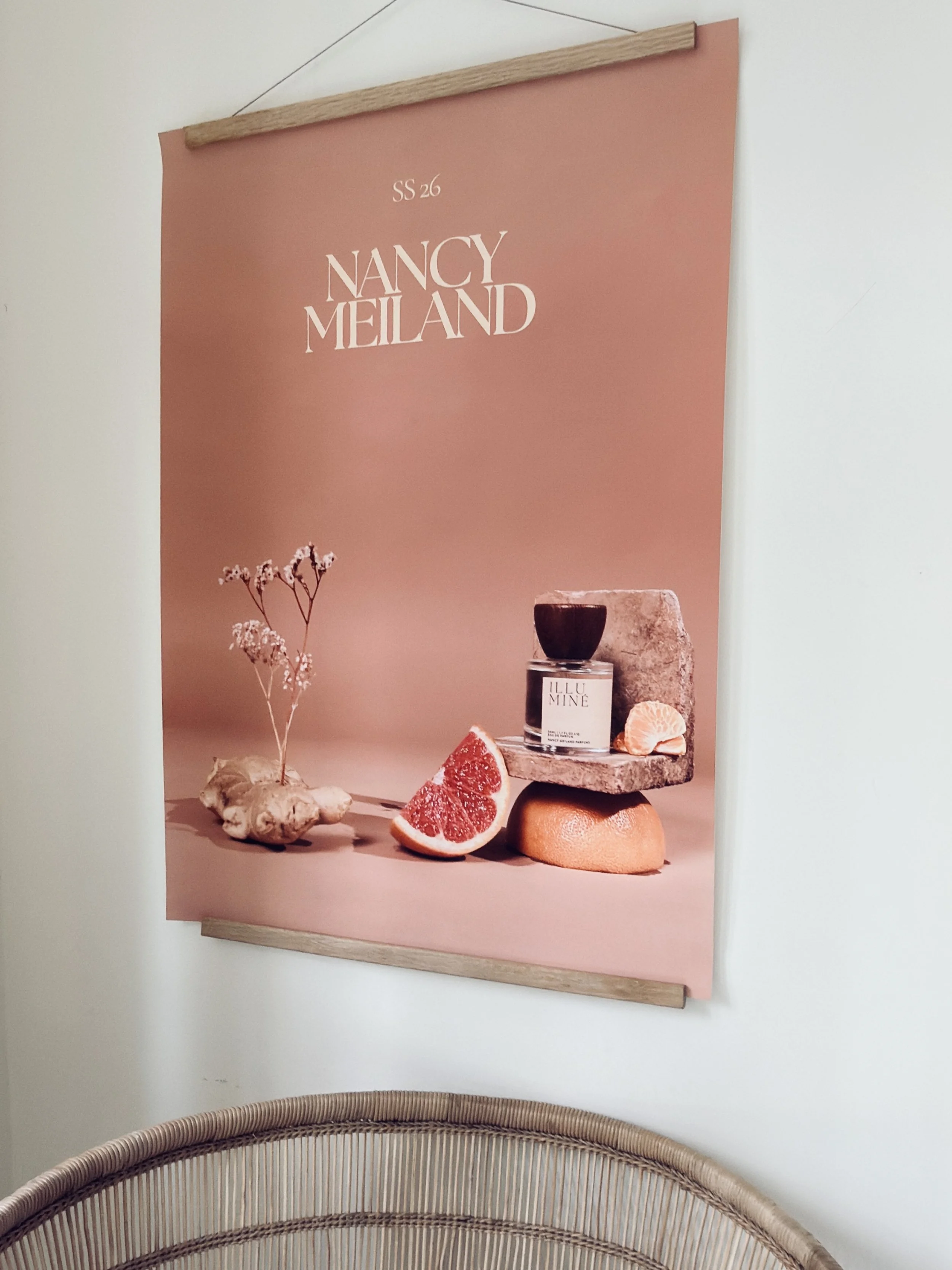 A poster with a pink background displaying the text "SS 26 NANCY MEILAND" alongside still life objects including a grapefruit, ginger root, a black candle in a glass container, a small gray stone, and a piece of clay or coral. The poster is hung on a