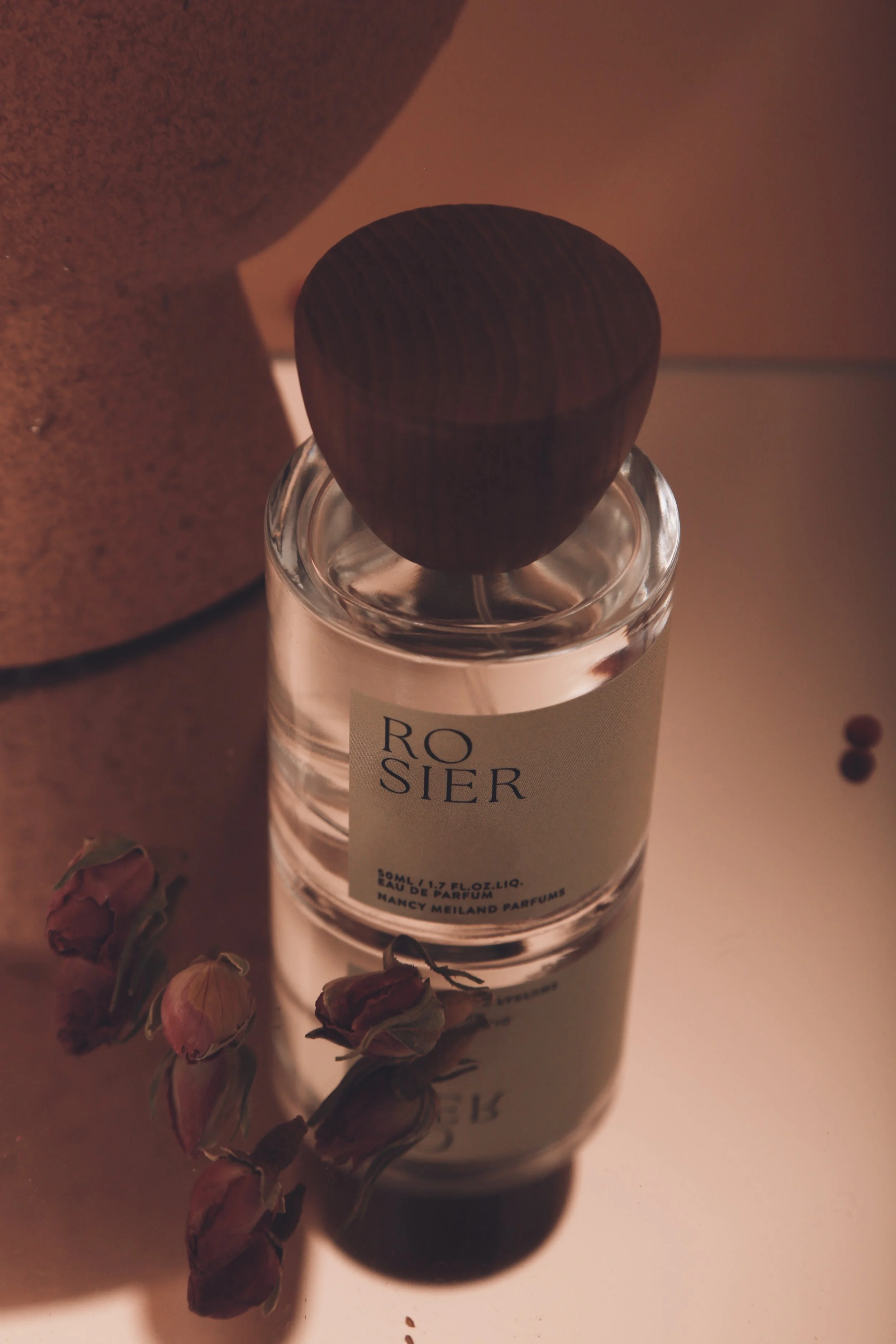 A bottle of perfume labeled 'Rosier' with a dark wooden cap, placed on a light-colored surface next to dried rose buds and a large cylindrical object, with warm, soft lighting.