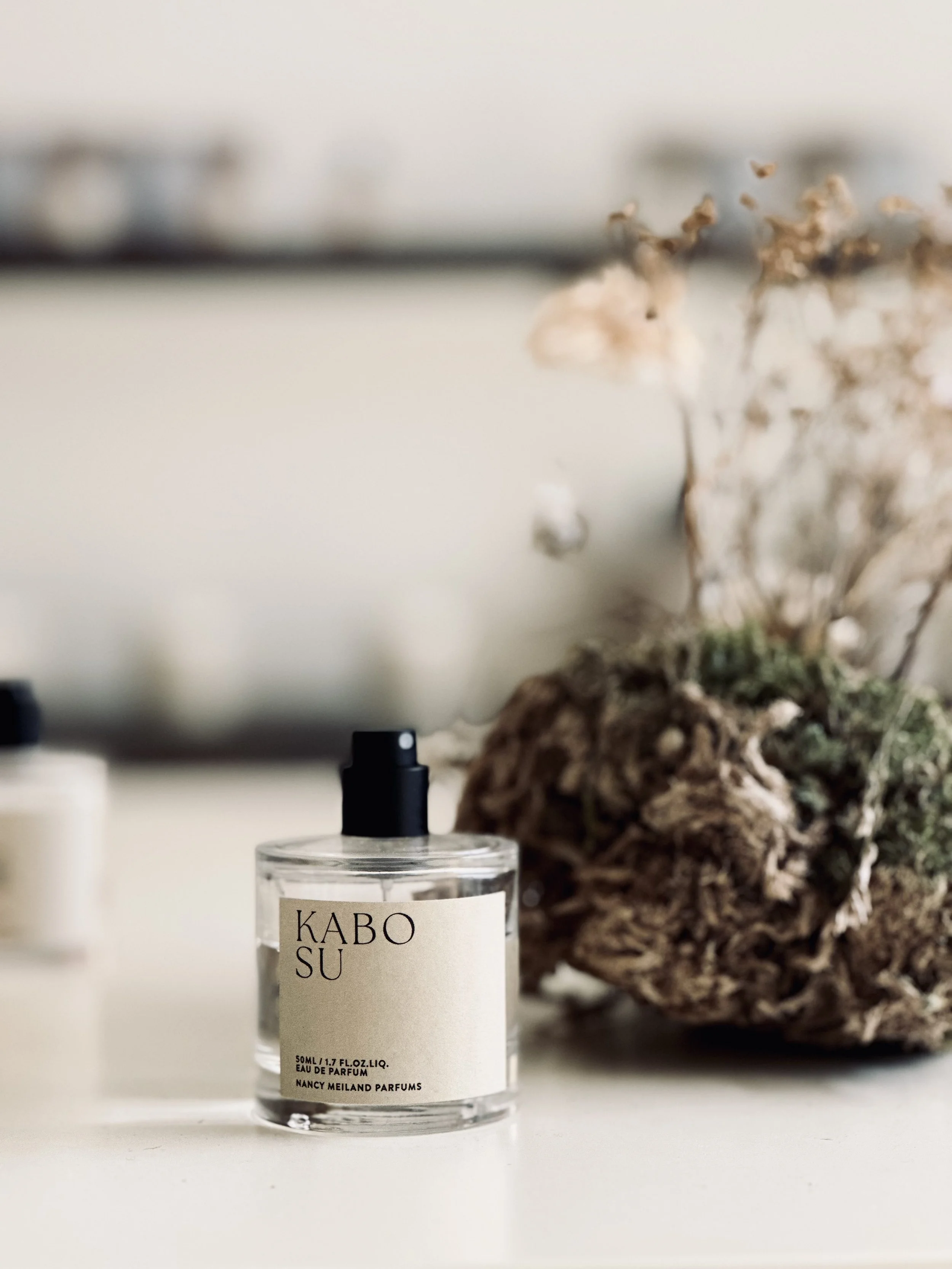 A clear glass bottle of perfume labeled 'KABO SU' with a black spray nozzle, placed on a white surface next to a dried plant arrangement.