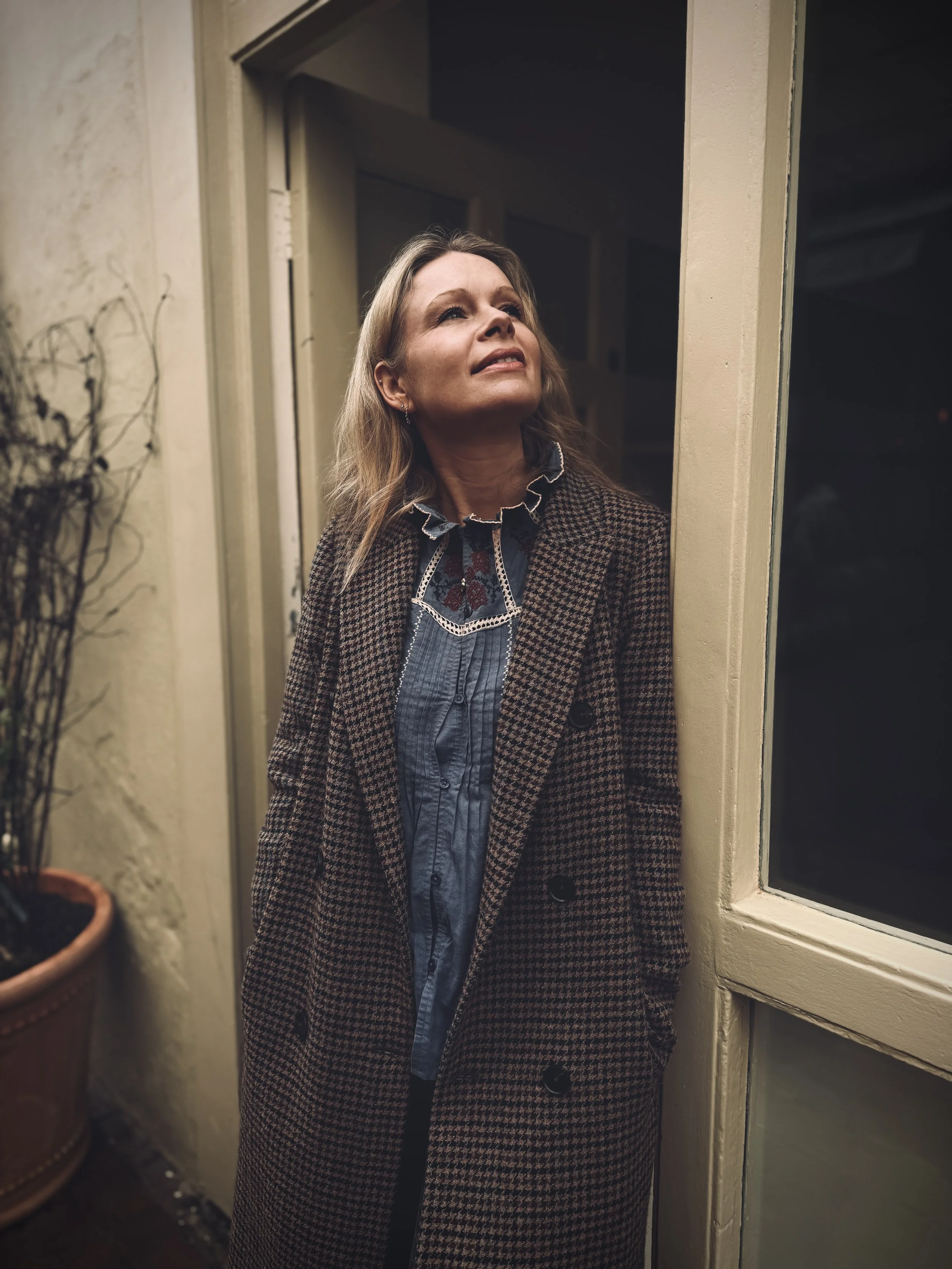 A woman with blonde hair wearing a checkered coat and a blue shirt stands at a door frame, looking upward with a concerned or pensive expression, inside a dimly lit room.