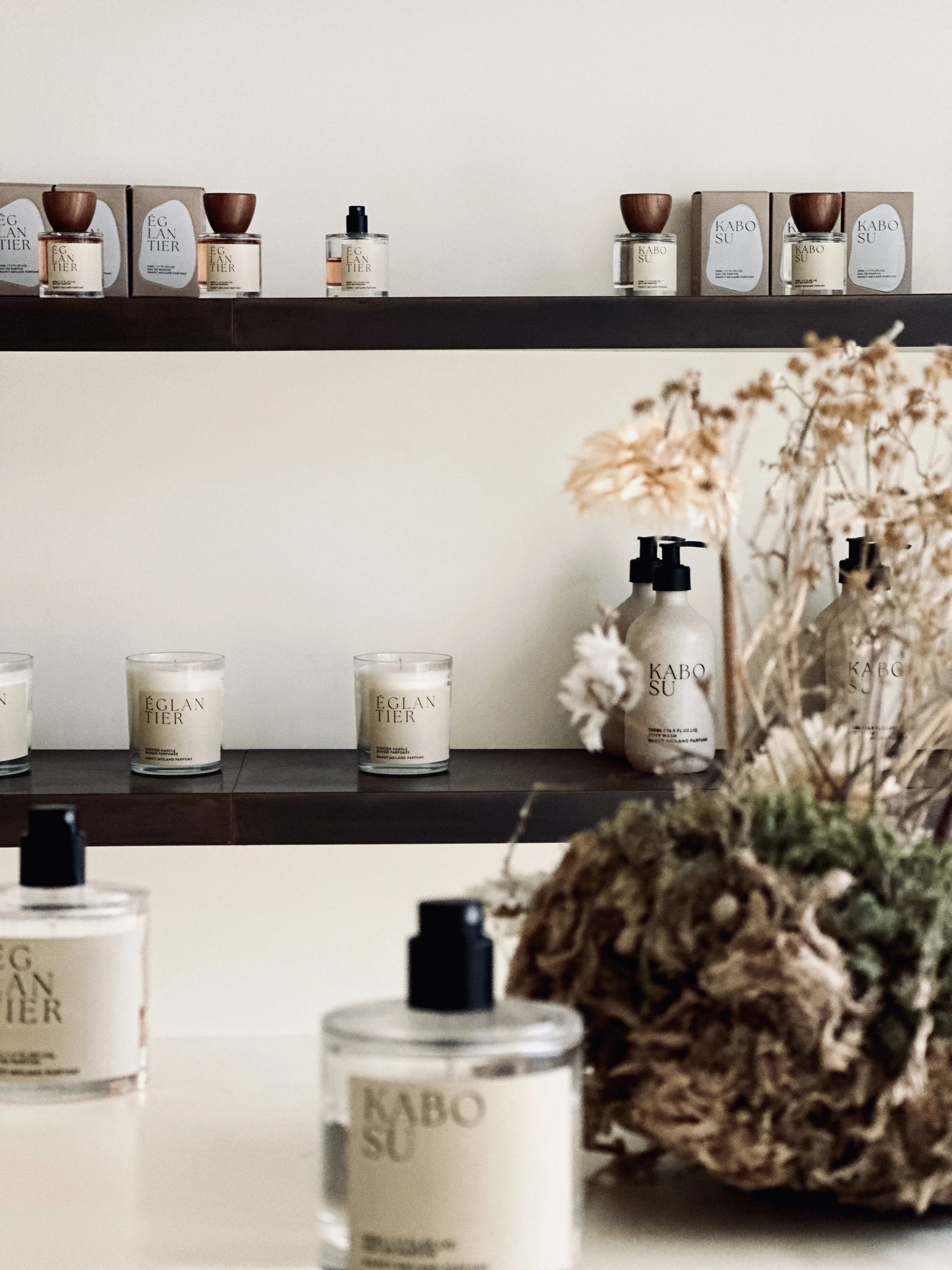 Display of luxury skincare and candle products on dark brown shelves, with decorative dried flowers in the foreground.