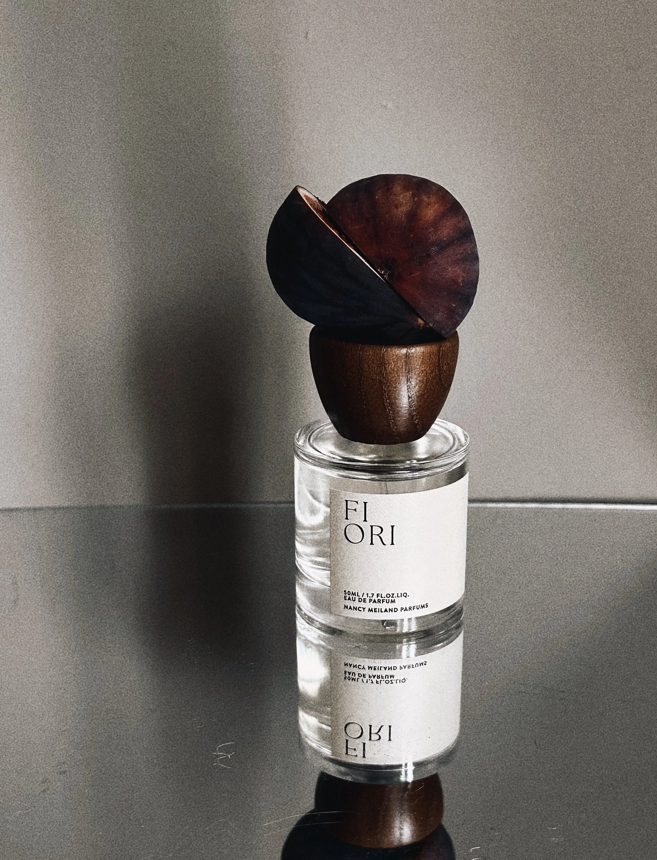 A perfume bottle with a minimalist label reading 'Fiori' and a wooden bowl with dark purple leaves balanced on top, reflected on a glossy surface.