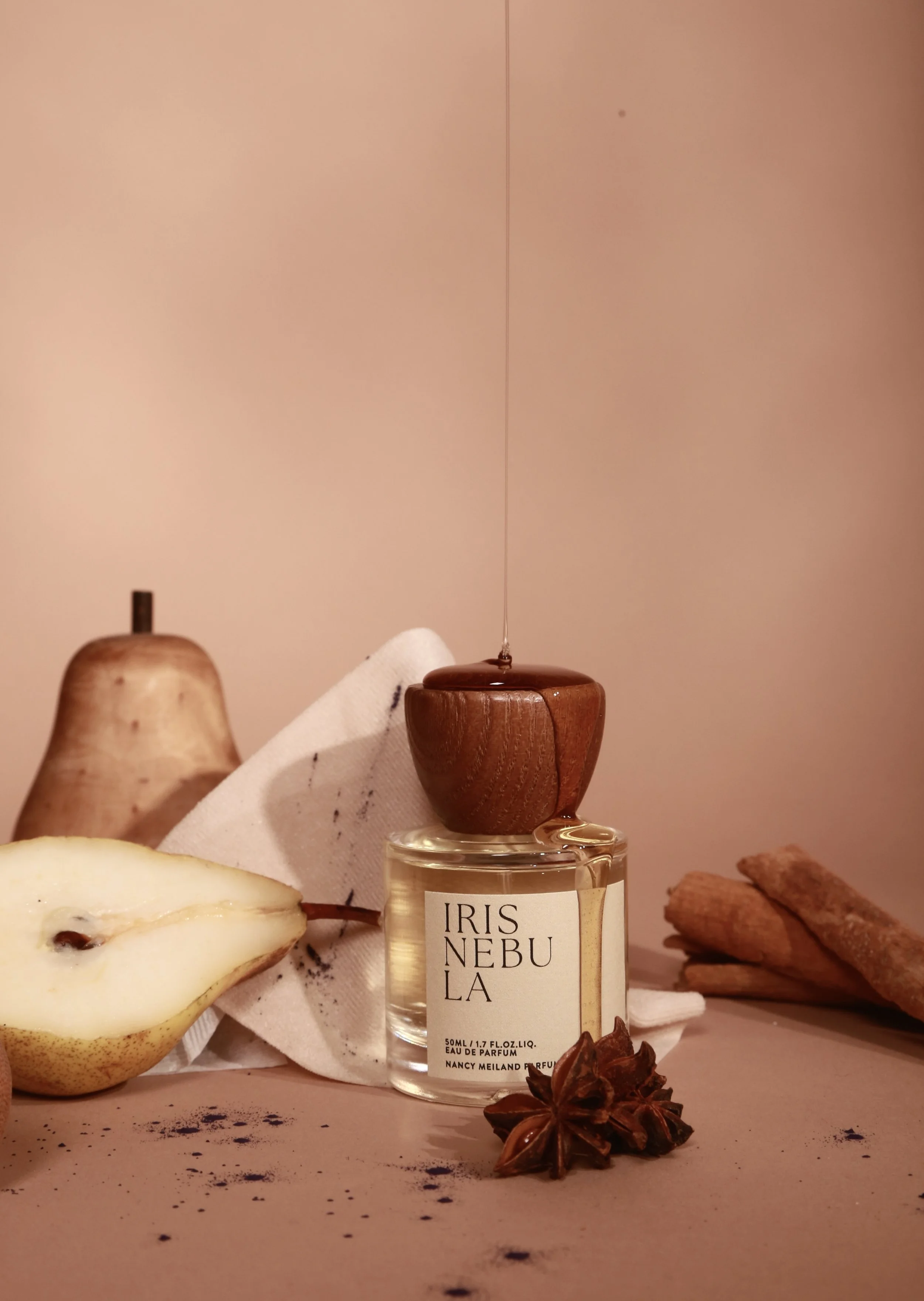 A bottle of Iris Nebula perfume with a wooden lid, surrounded by a halved pear, cinnamon sticks, star anise, and a piece of white cloth against a pinkish background.