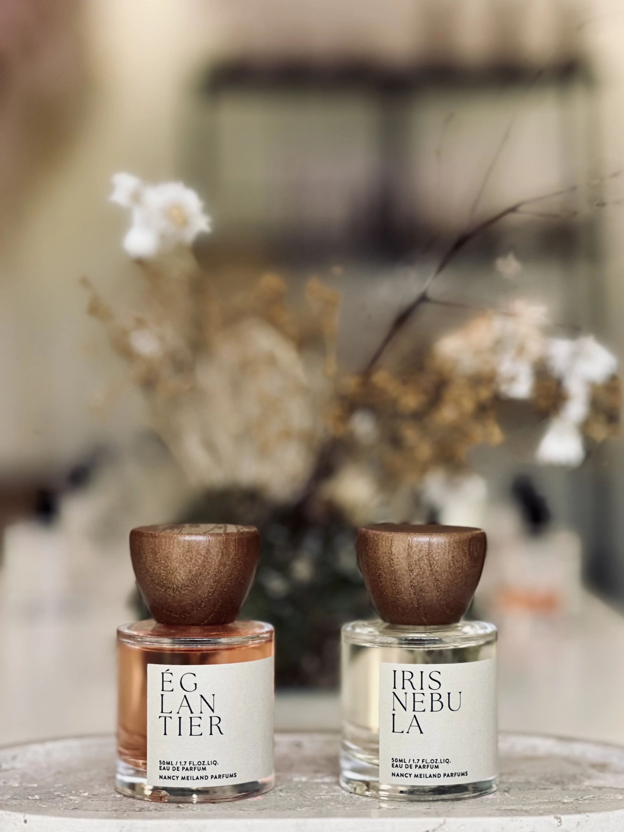 Two small perfume bottles with wooden caps, labeled 'ÉCLANTIER' and 'IRIS NEBULA,' placed on a surface with a blurred floral background.