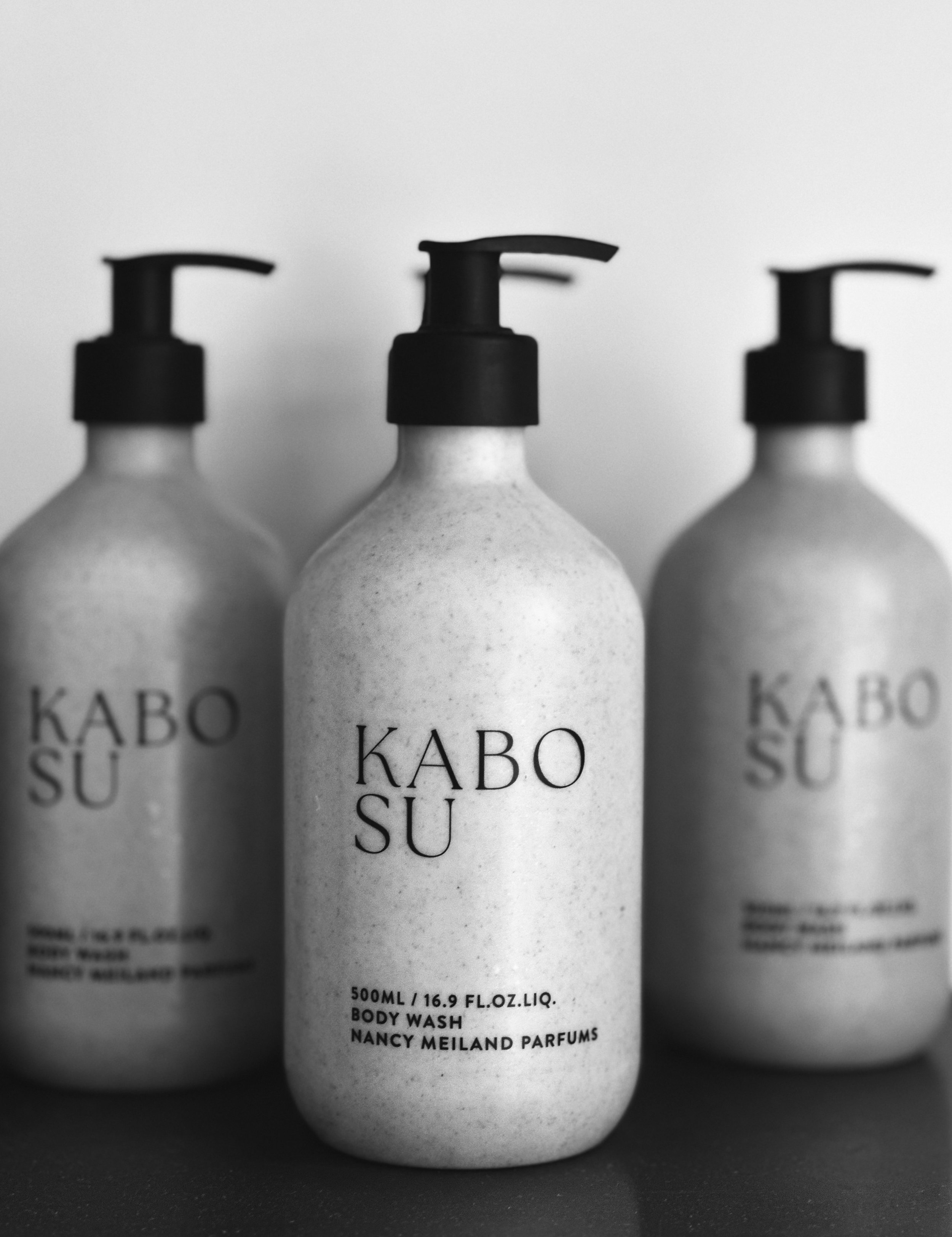 Three bottles of KABO SU body wash, with the middle bottle in focus, black pump dispensers, against a plain background, in black and white.