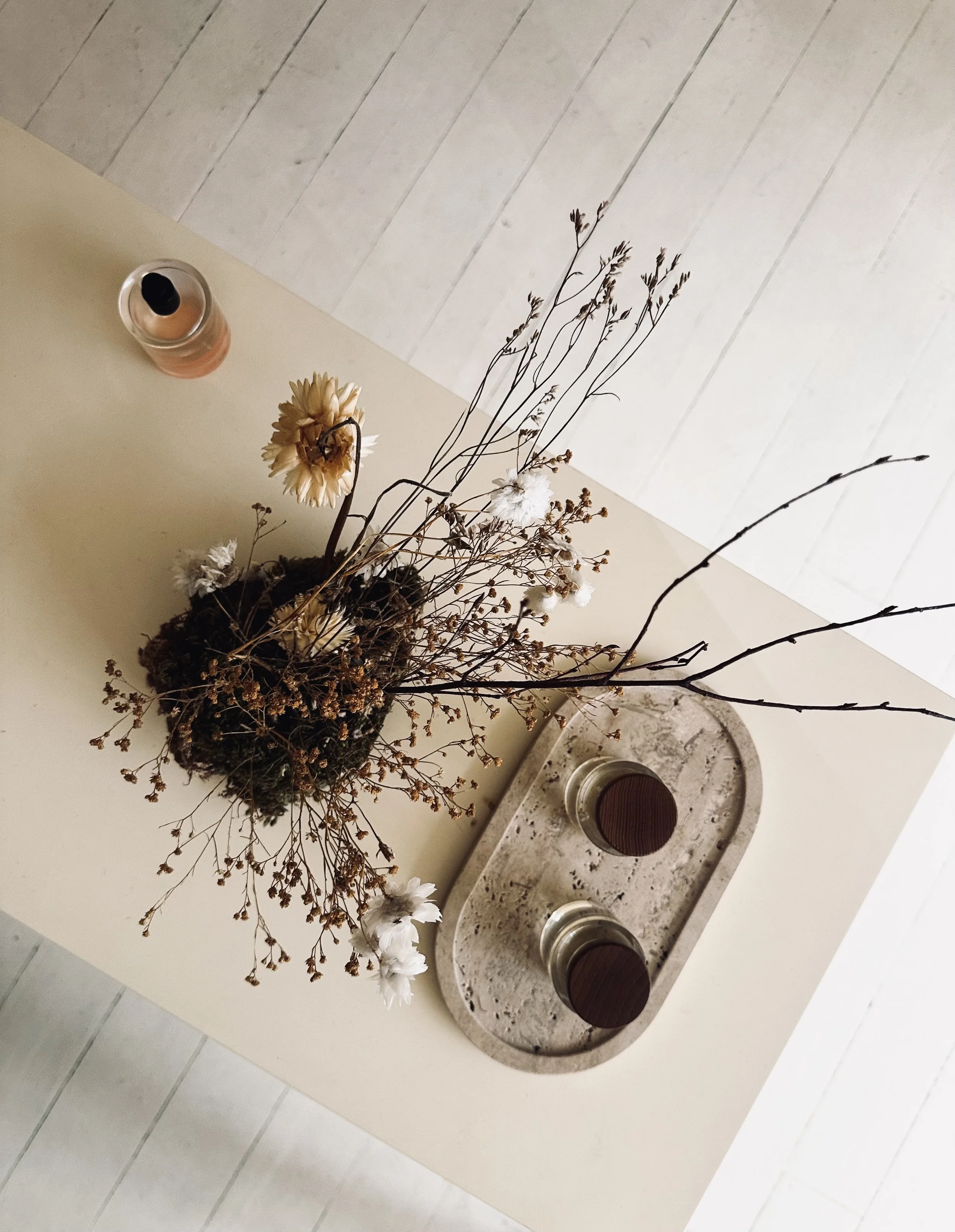 A beige table with a dried floral arrangement in a dark round vase, two candles in glass holders, a tray with two candles, and a light-colored wooden floor in the background.
