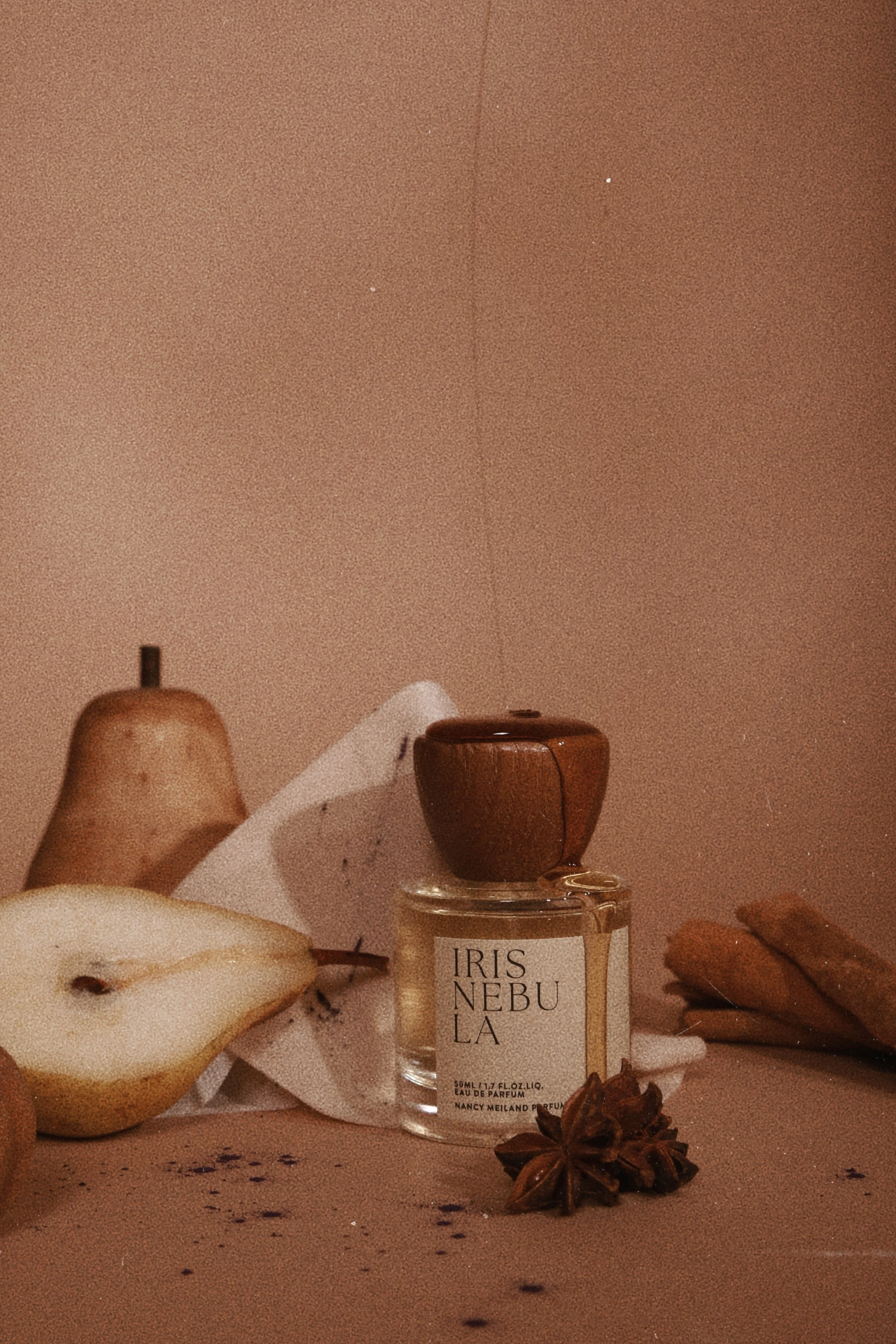 A still life composition featuring a perfume bottle labeled 'IRIS NEBULA' with a brown cap, a halved pear, a star anise, and a small pear on a peach-colored surface and background, with a white cloth and some cookies in the background.