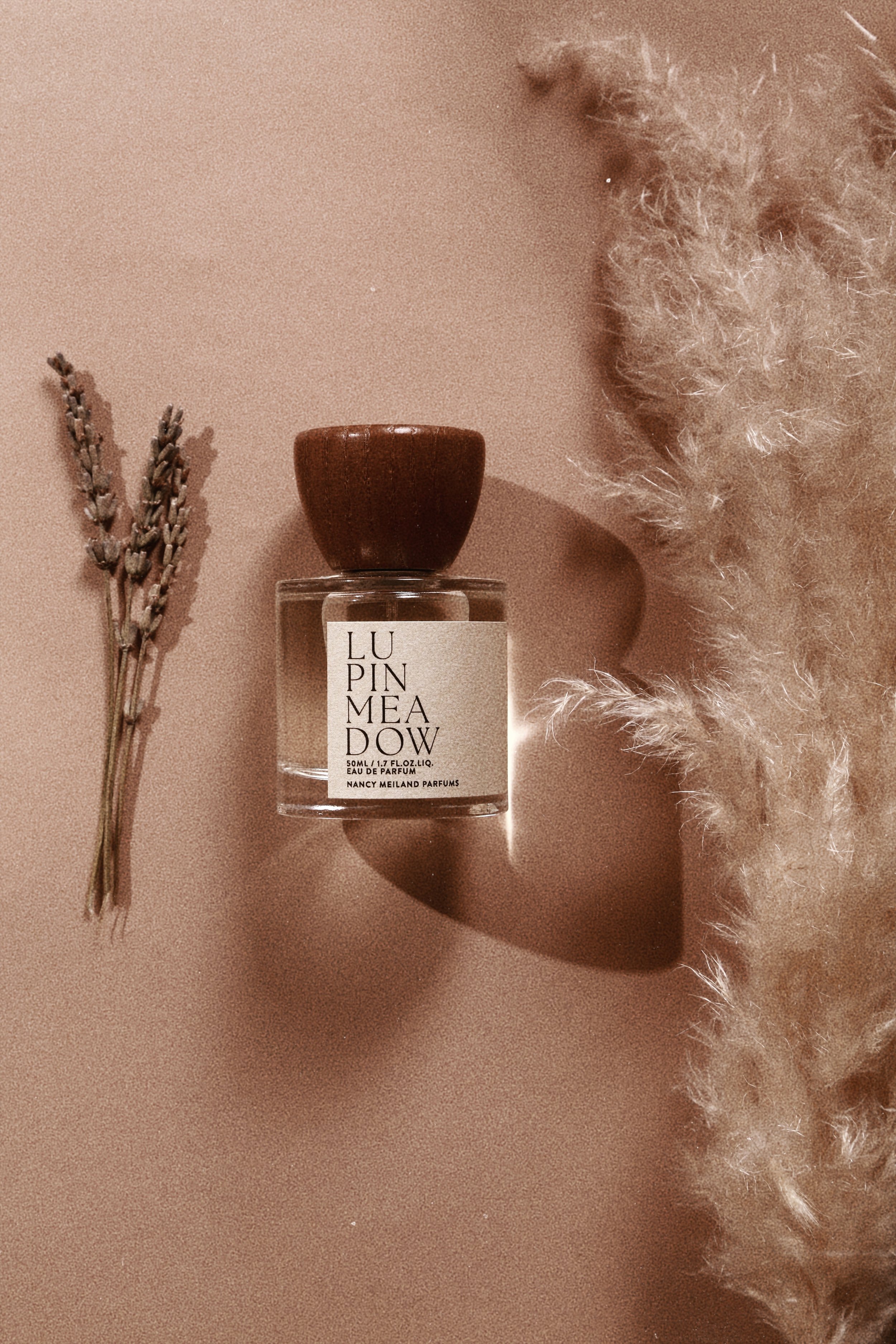 A perfume bottle labeled 'LUMI MEADOW' with a wooden cap, placed on a pink surface alongside a small bouquet of lavender and a fluffy beige fabric.