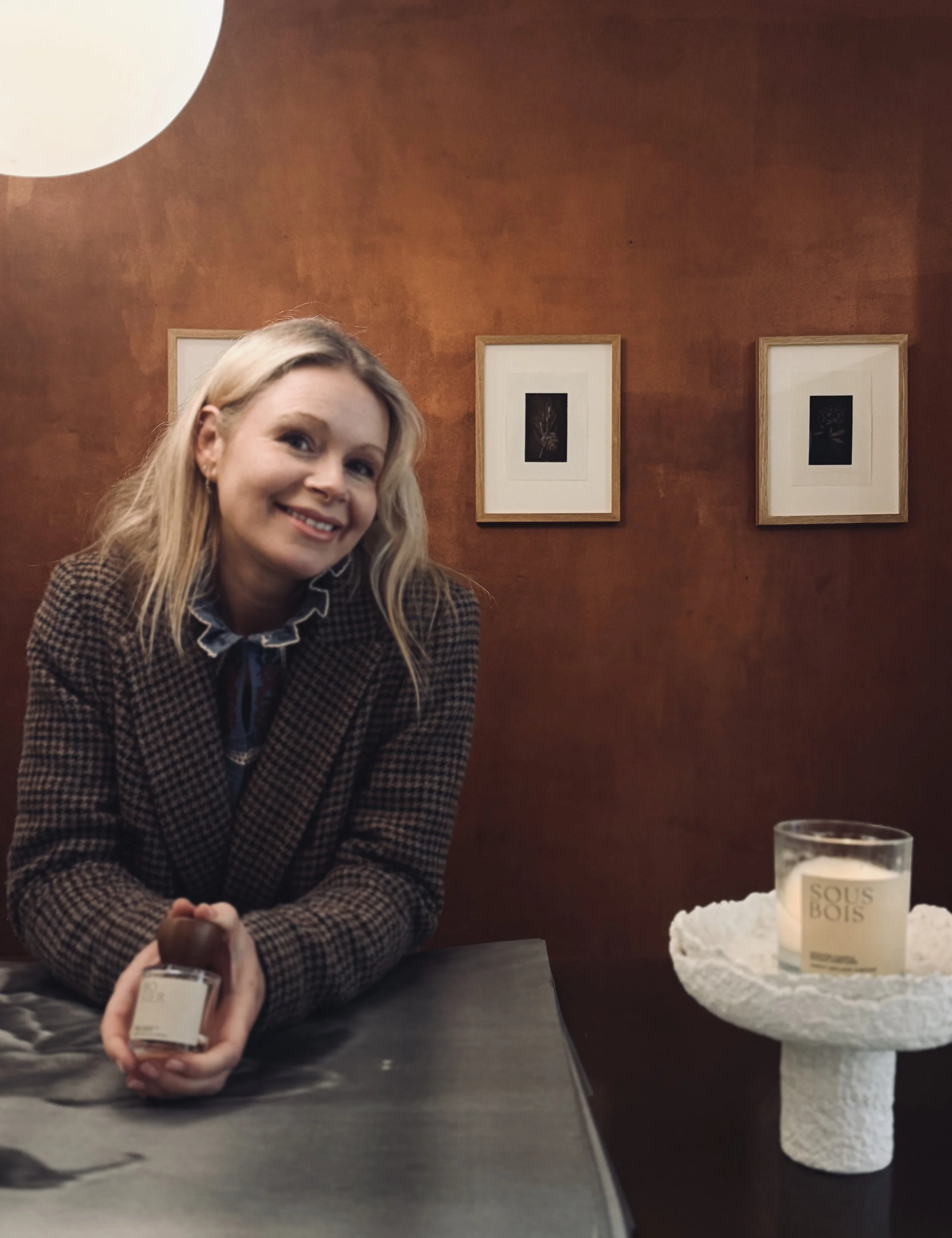 A smiling woman with blonde hair wearing a brown checkered blazer sits at a table in a warmly lit room with a brown wall decorated with three framed artworks. She holds a small glass bottle in her hands, and on the table there is a white textured can