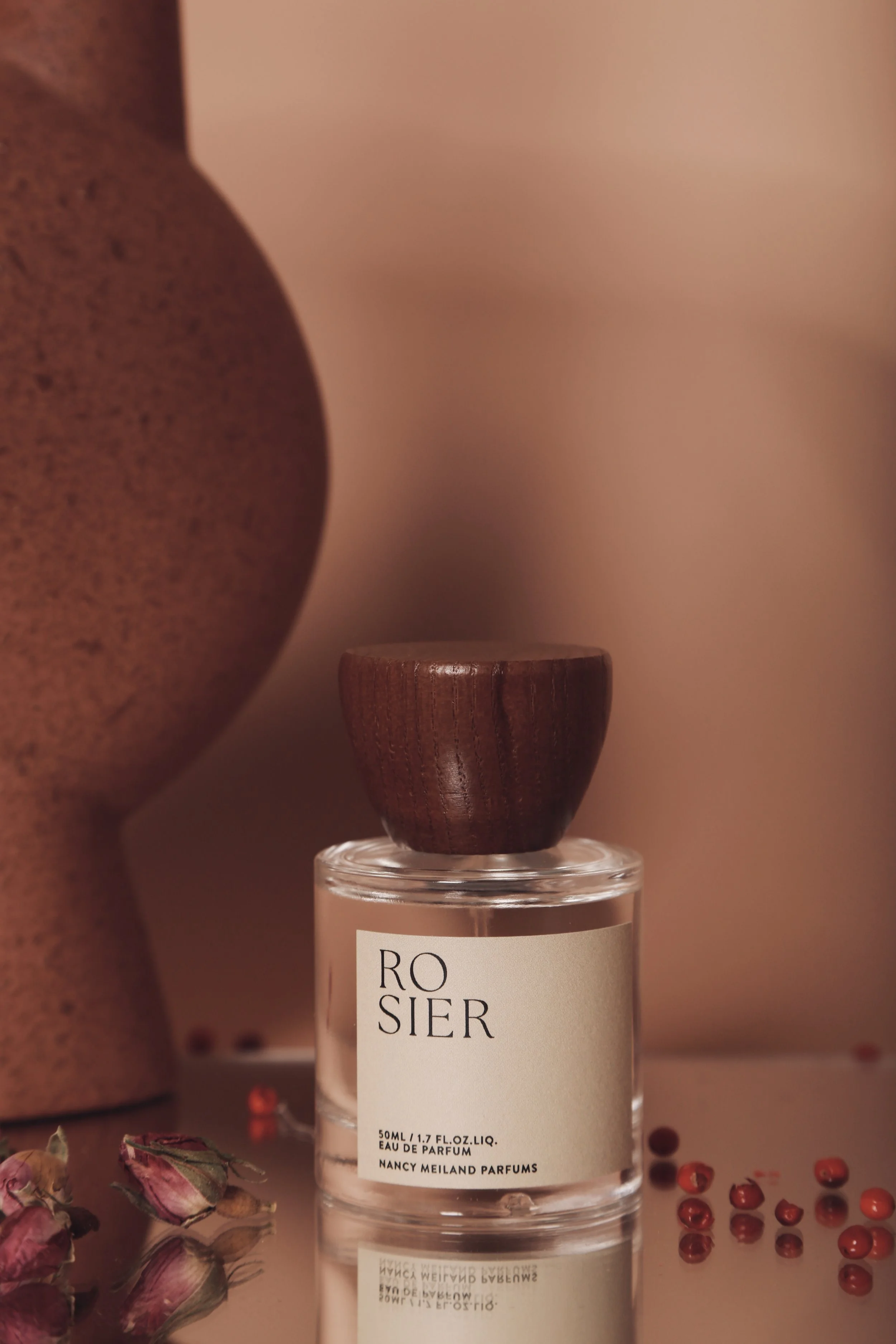 A perfume bottle labeled 'Rosier' with a wooden cap, placed on a reflective surface, surrounded by dried roses and red peppercorns, with a blurred ceramic vase in the background.