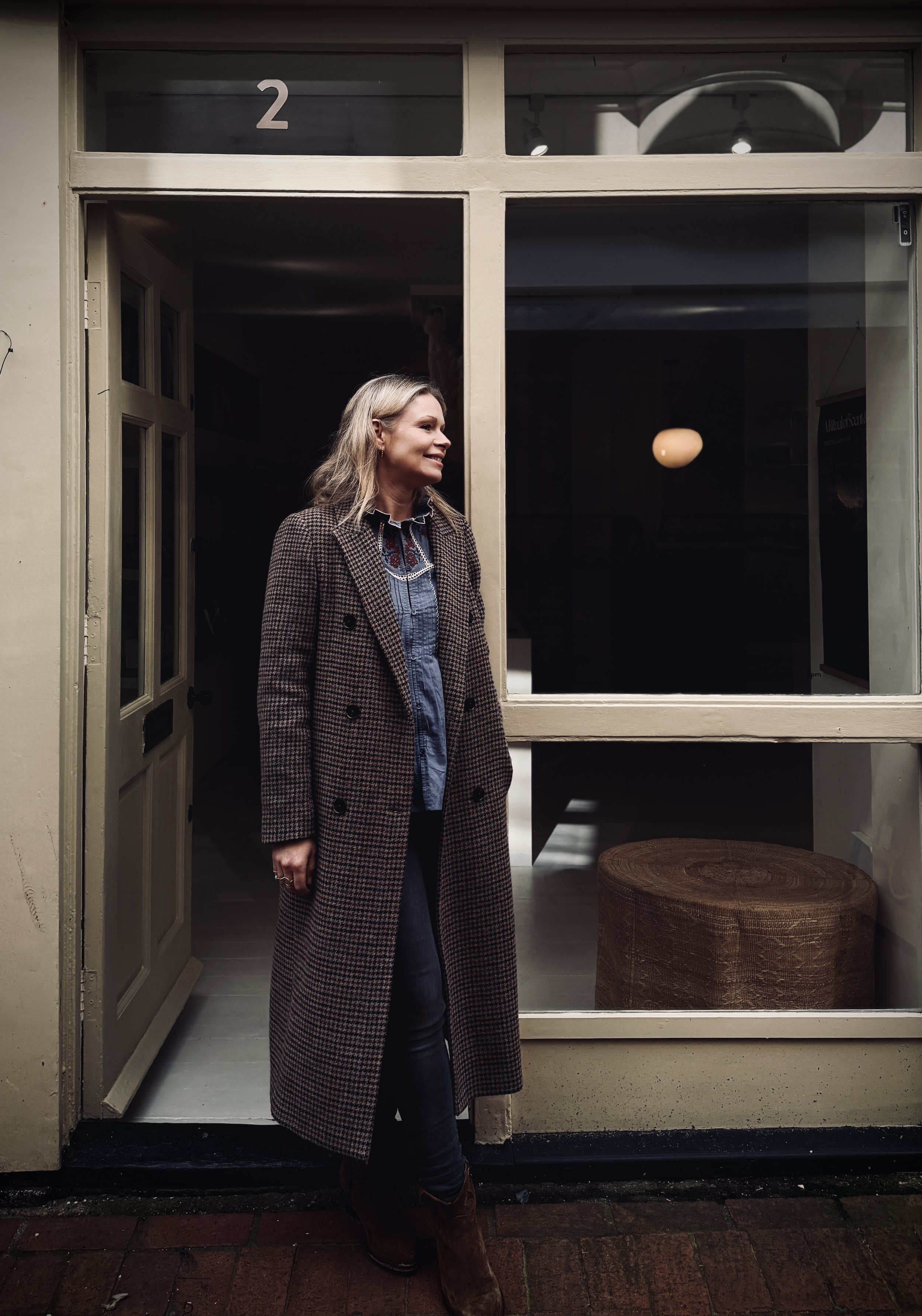A woman standing in front of a storefront window with the number 2 on it, wearing a long checkered coat and smiling.
