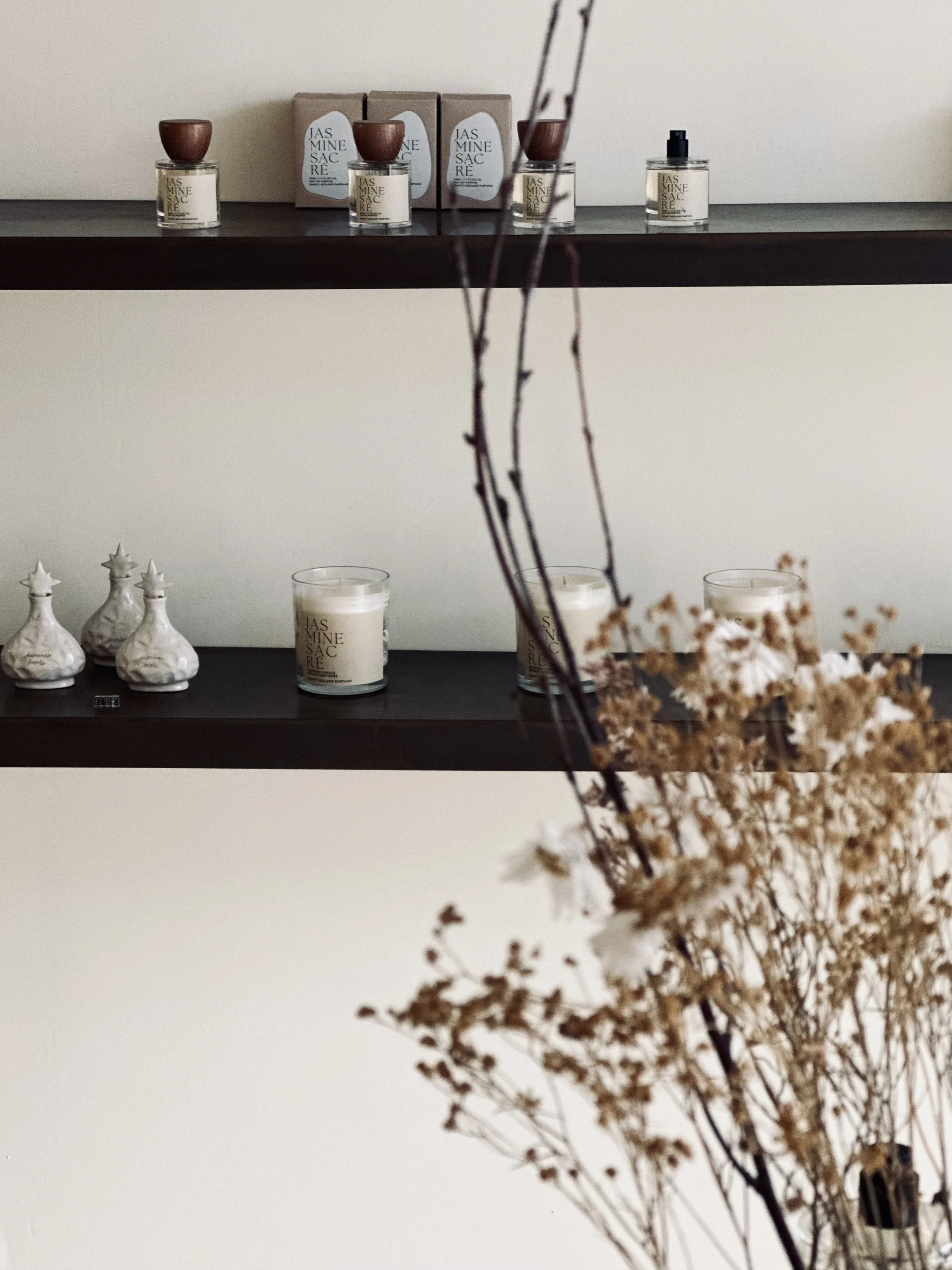 Decorative shelves displaying candles, bottles, and ceramic figurines, with dried branches and flowers in the foreground.