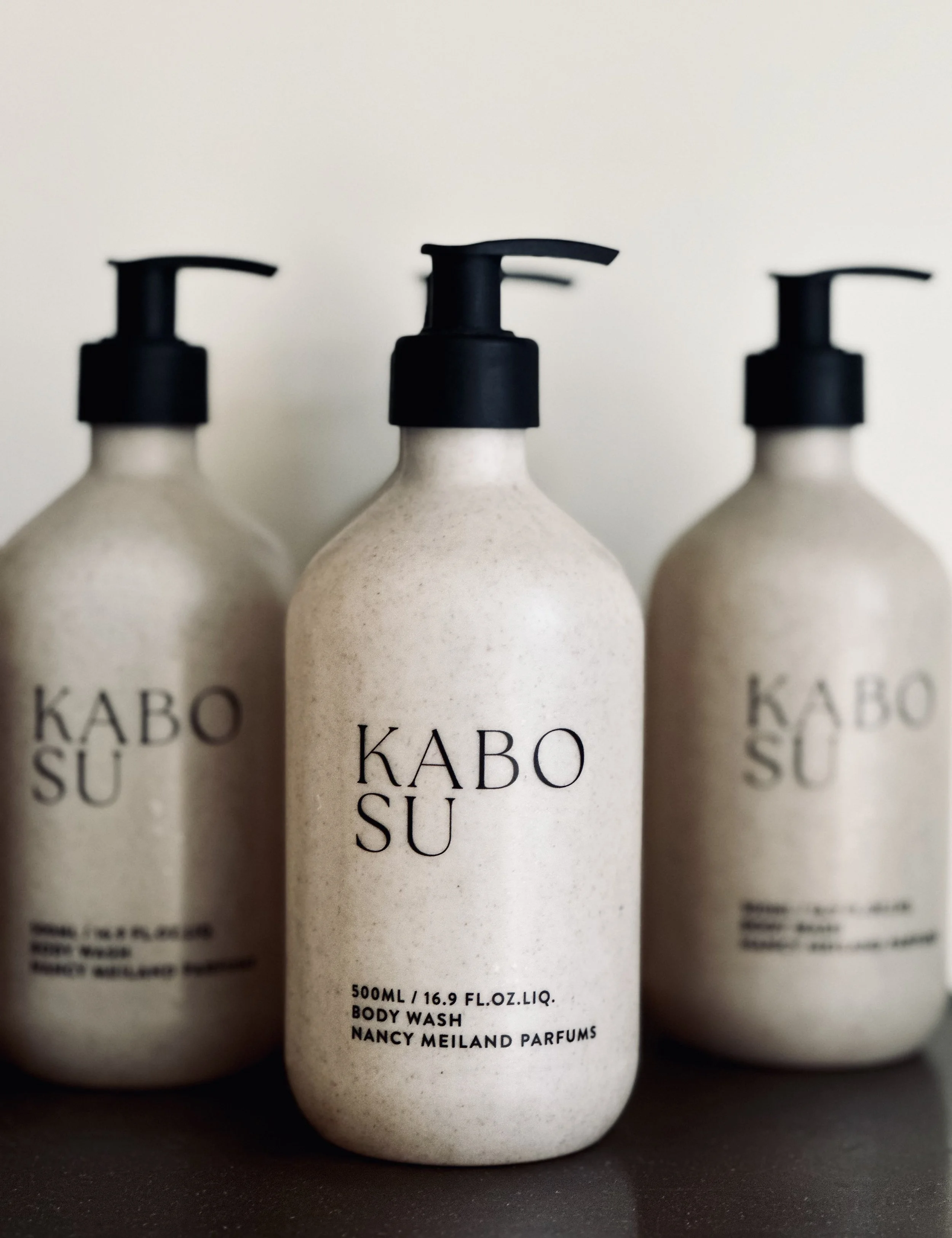 Three beige bottles with black pump dispensers labeled "KABO SU," with the front bottle identified as body wash from Nancy Meiland Parfums.