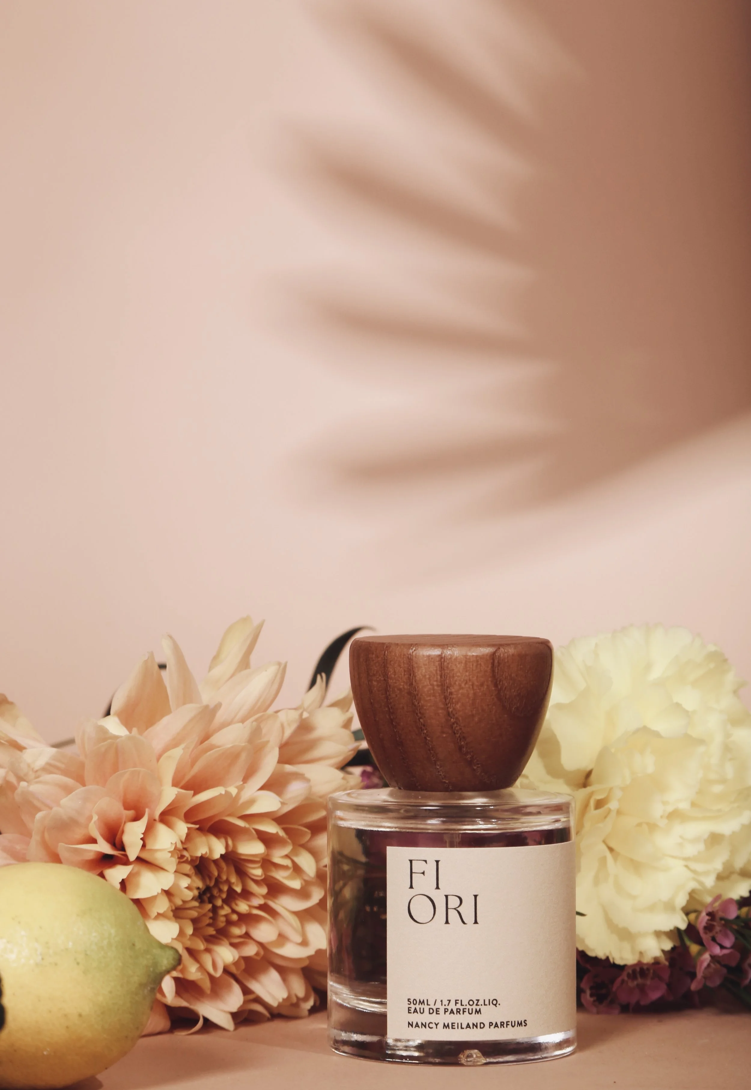 A bottle of Nancy Meiland Parfums Eau de Parfum labeled Flori with a wooden cap, surrounded by light pink and cream flowers, a lemon, and a soft pink background with a blurred palm leaf shadow.