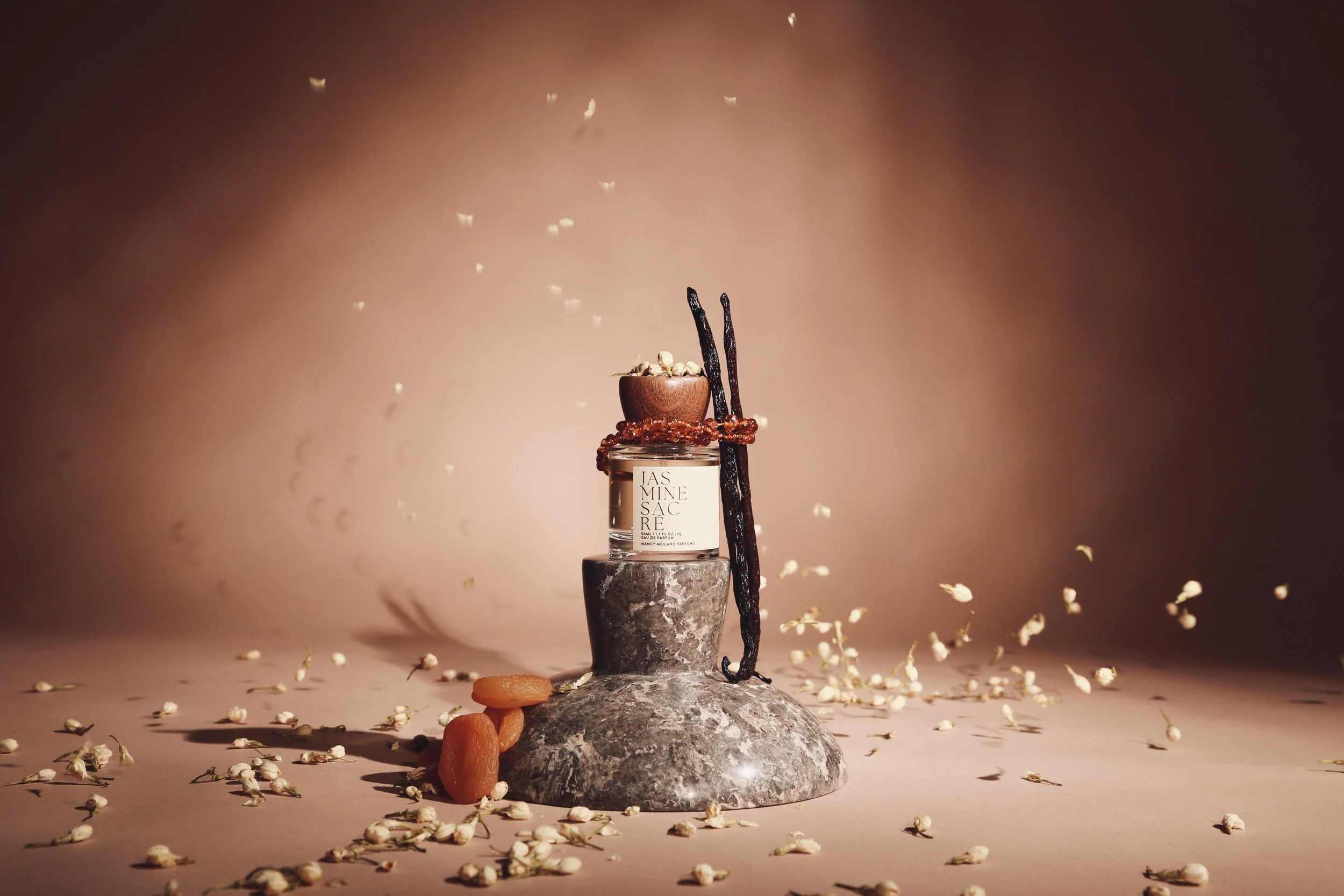 A perfume bottle labeled 'Jasmine Sacré' is placed on a stone pedestal against a peach-colored background, decorated with dried apricots, vanilla beans, a small bowl of jasmine flowers, and scattered jasmine petals.