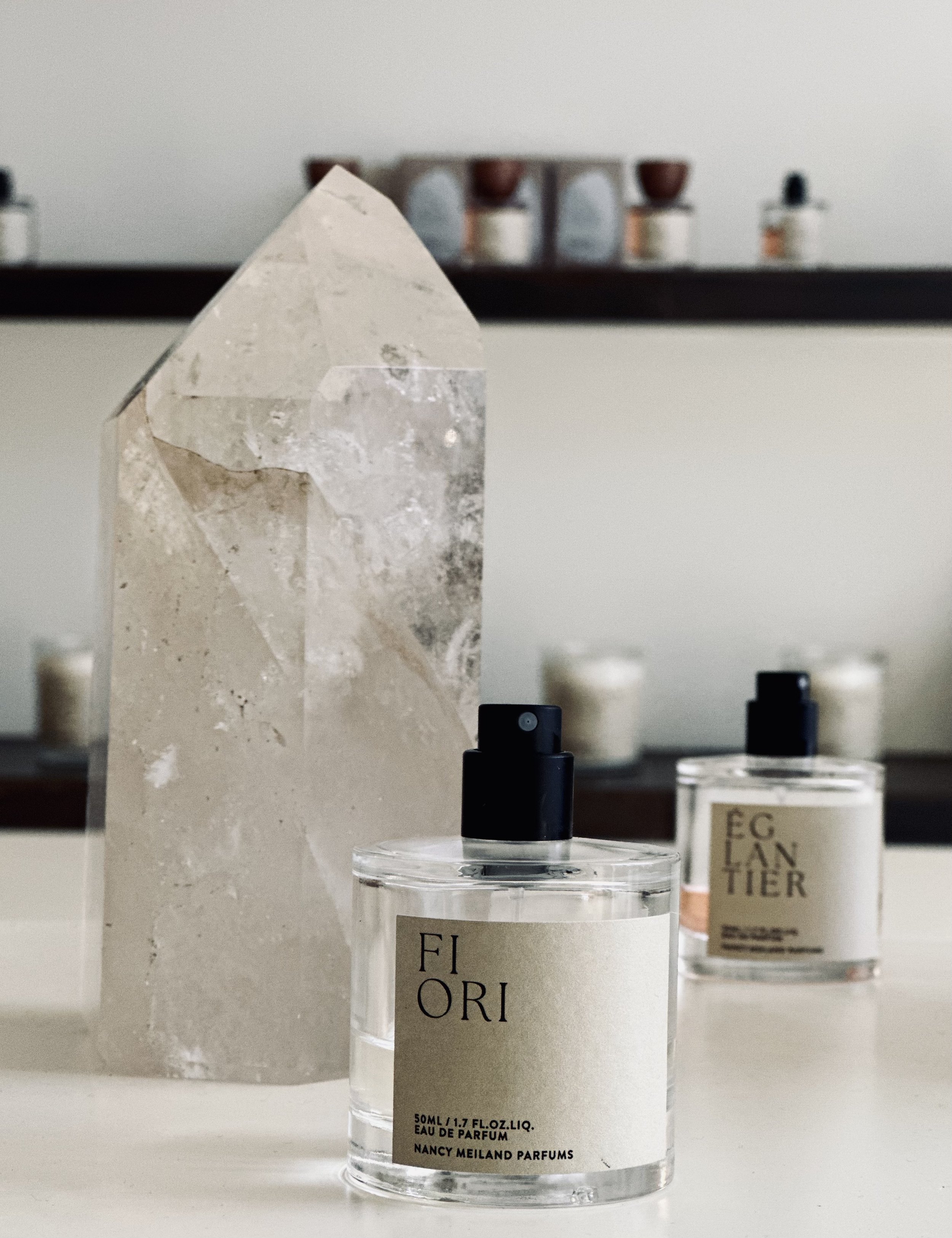 Two bottles of Nancy Meiland Parfums' Fior perfume on a white surface with a large, clear, geometric-shaped crystal and a black shelf with small jars in the background.