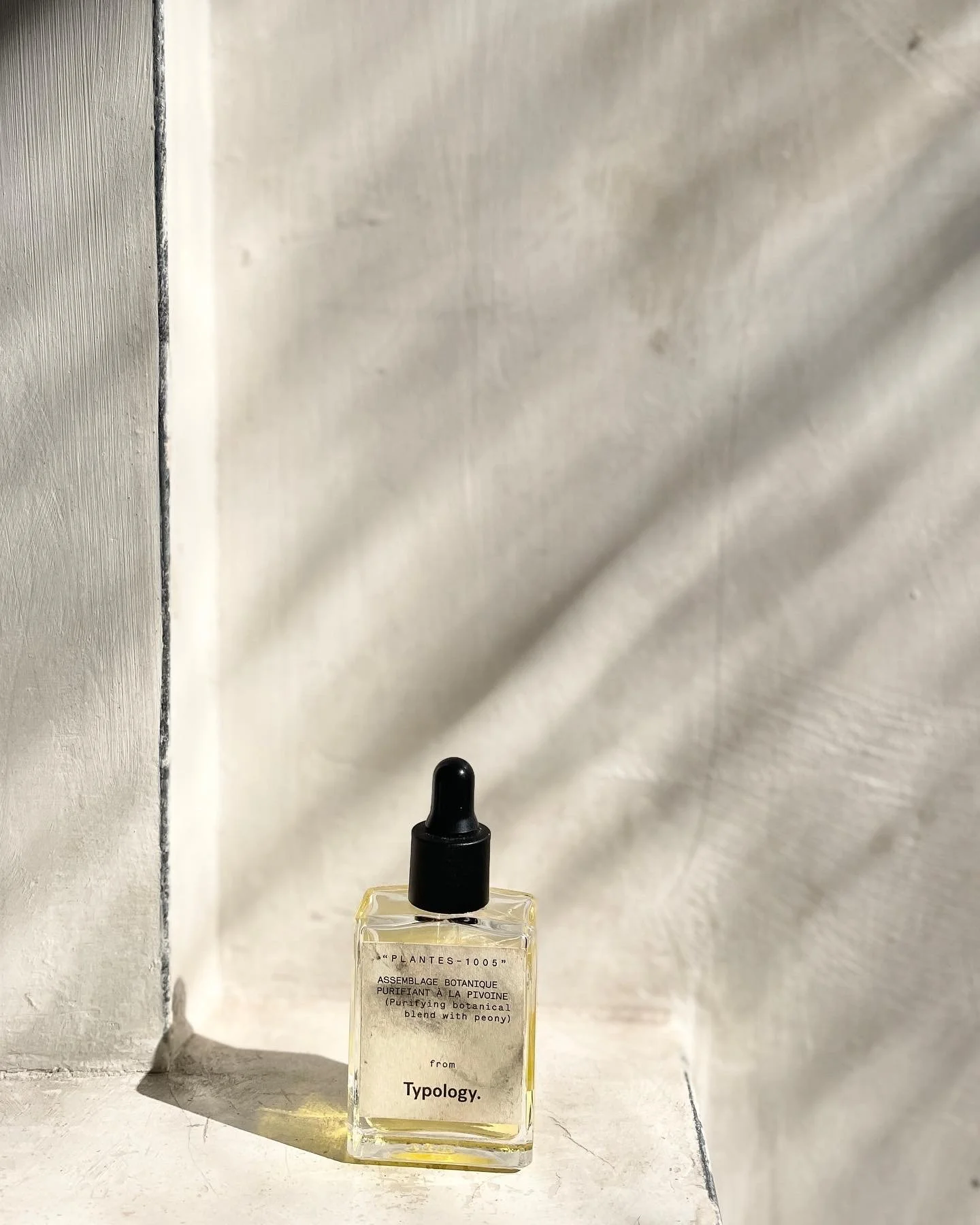 A small glass bottle with a black dropper cap, containing a light yellow liquid, placed on a white surface with a shadow cast behind it. The bottle has text on it that reads 'from Typology.' and some additional smaller text.