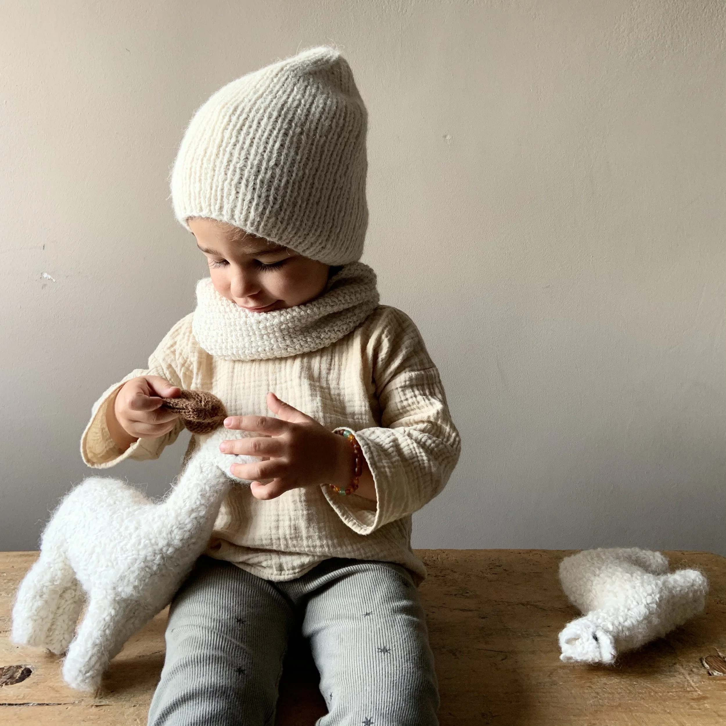 A young child wearing a beige knit hat and scarf, sitting at a wooden table, holding a white knitted sock and a small brown sock, with another white sock on the table beside them.