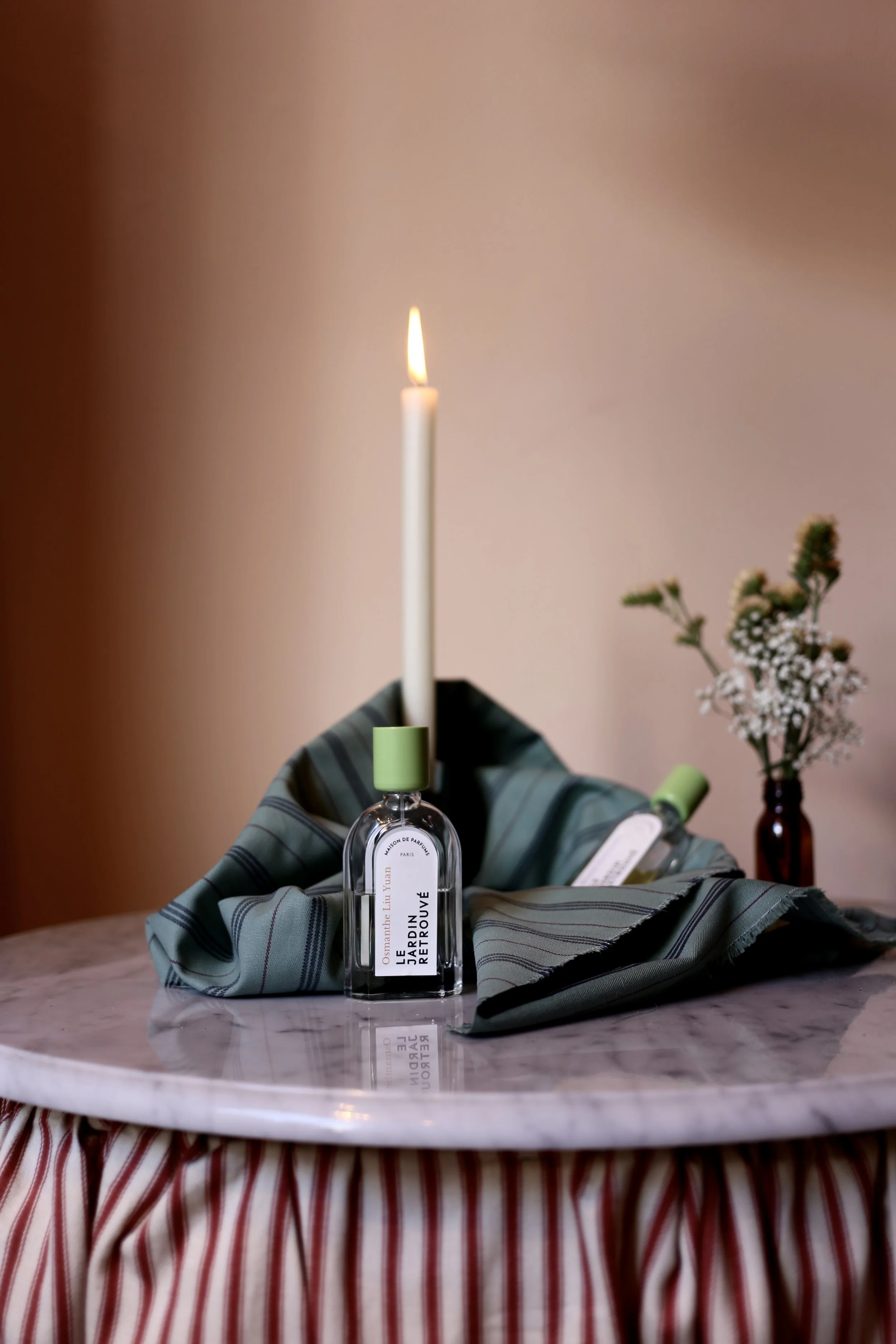 A marble-topped table with a striped tablecloth, a lit candle in a perfume bottle, and a small vase with flowers in the background.
