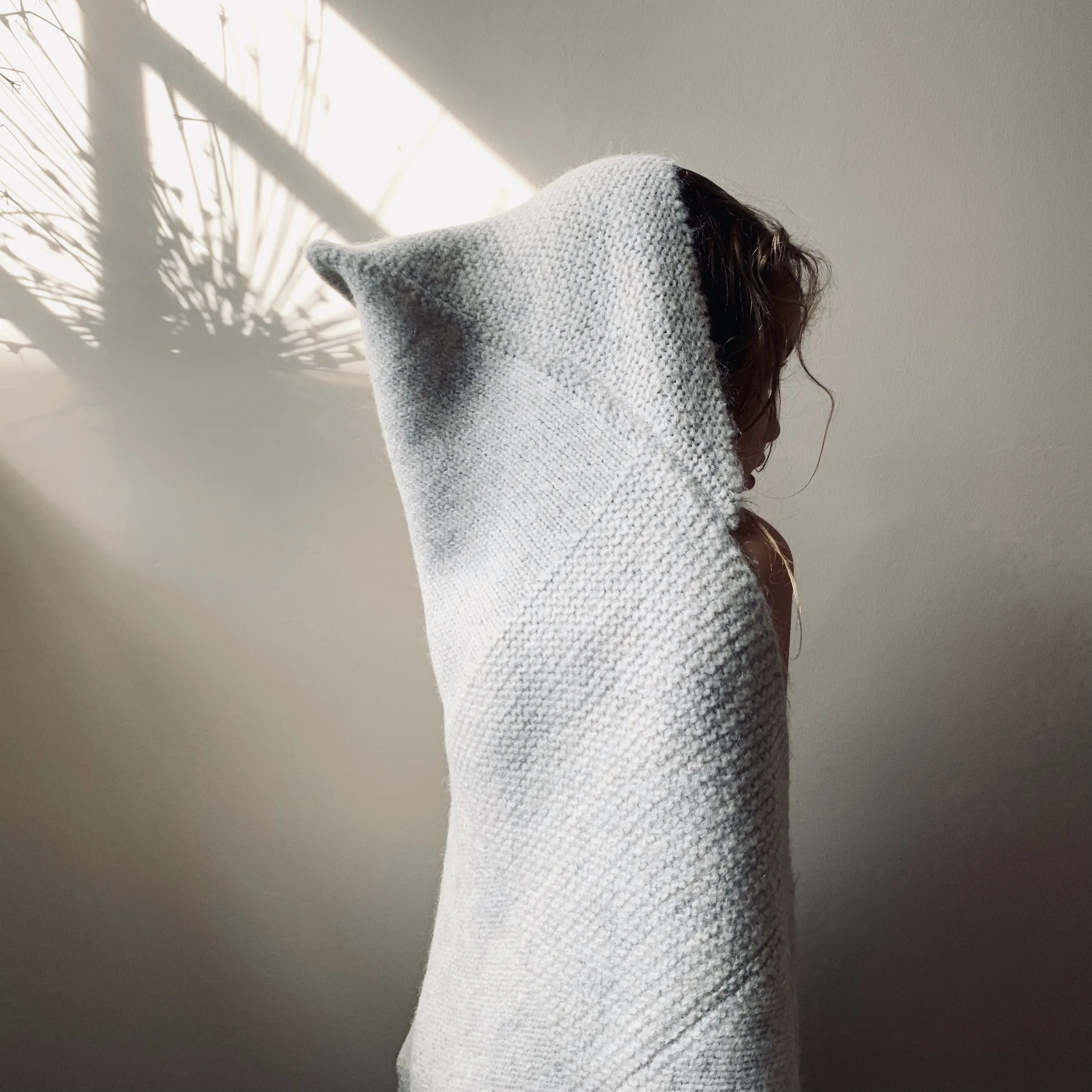 Person with brown hair partially covered by a gray knit hoodie standing against a light-colored wall with shadow of a plant cast on it.